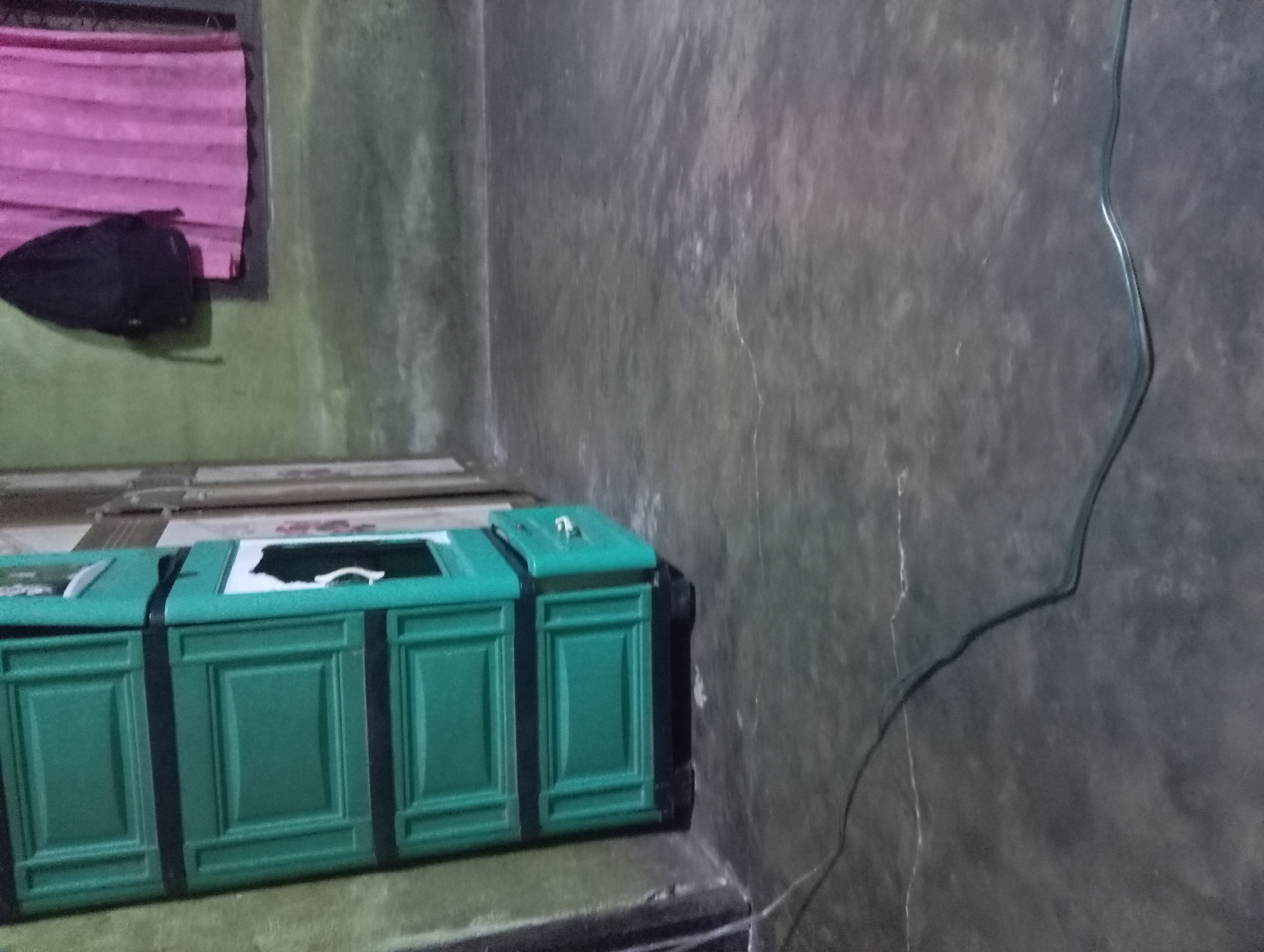 In a room in Rejosari, Indonesia, a tall, green plastic cabinet with three drawers stands against a green wall. The cabinet has black trim and the top drawer is slightly ajar. To the right of the cabinet, a pink curtain hangs over a window. A black backpack is suspended from the wall above the window. The floor is concrete and shows some cracks. A dark electrical cord snakes across the floor in the foreground.