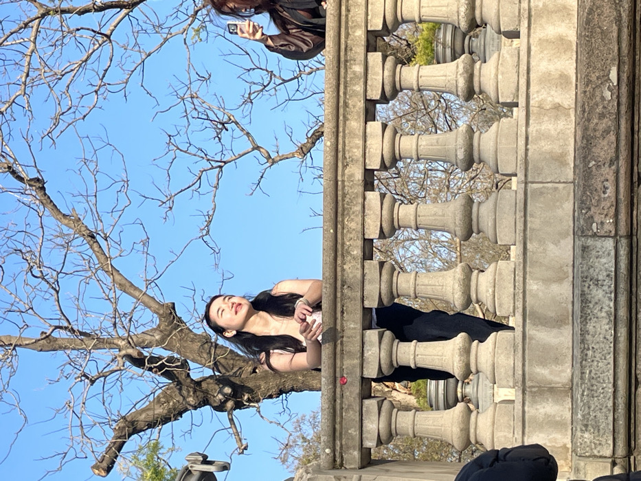 The image shows two individuals interacting on an elevated stone balustrade within Parc de la Ciutadella, Barcelona, Spain. The scene is set against a clear blue sky, with several large, bare deciduous trees forming the background above the balustrade.
In the center-left, a young East Asian female with long dark hair stands partially obscured by the balustrade, from the waist up. She is wearing a light-colored sleeveless top and dark pants. Her head is tilted slightly upward and to the right, with her eyes closed, as if experiencing sunlight. Her left arm rests on the balustrade, and she holds a small white object, possibly a cup or phone.
To her right, partially visible above the balustrade, is a second individual with dark, wavy hair, wearing a dark brown or olive-colored jacket. This person is holding a white smartphone horizontally with their right hand, pointing it towards the left, ostensibly recording or photographing the first individual or the general scene.
The prominent foreground feature is a robust stone balustrade, composed of multiple light grey balusters and a flat top rail. A small red object, possibly a flower petal, rests on the top rail near the center-left. In the extreme bottom-left corner, a dark fabric garment, likely belonging to a third person, is partially visible. The lighting suggests bright, direct sunlight, illuminating the subjects and casting soft shadows. The leafless trees indicate a cool-season setting.