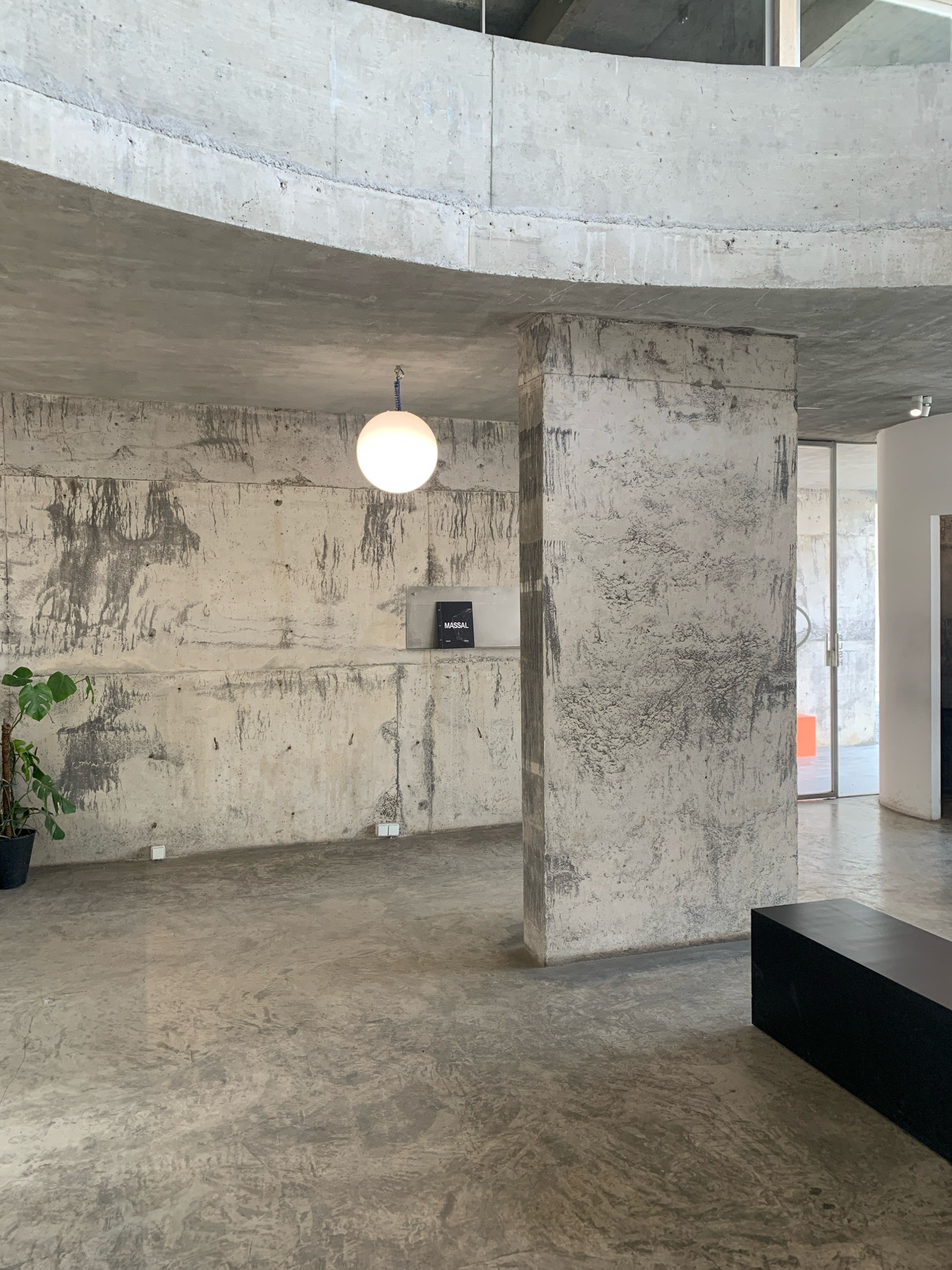 The image depicts the interior of a minimalist, brutalist-style space with raw concrete walls, floors, and ceilings. A large, square concrete pillar stands prominently in the foreground, its surface textured and stained. A spherical pendant light hangs from the ceiling, casting a warm glow. On the wall, a small, black book with the text "MASSAL" is displayed. To the left, a potted plant adds a touch of greenery. In the background, glass doors lead to another area, revealing glimpses of an orange object. A dark, rectangular bench is positioned in the lower right corner of the frame. The overall aesthetic is stark, industrial, and modern. This location is identified as MASSAL in the city of Meri, Indonesia.