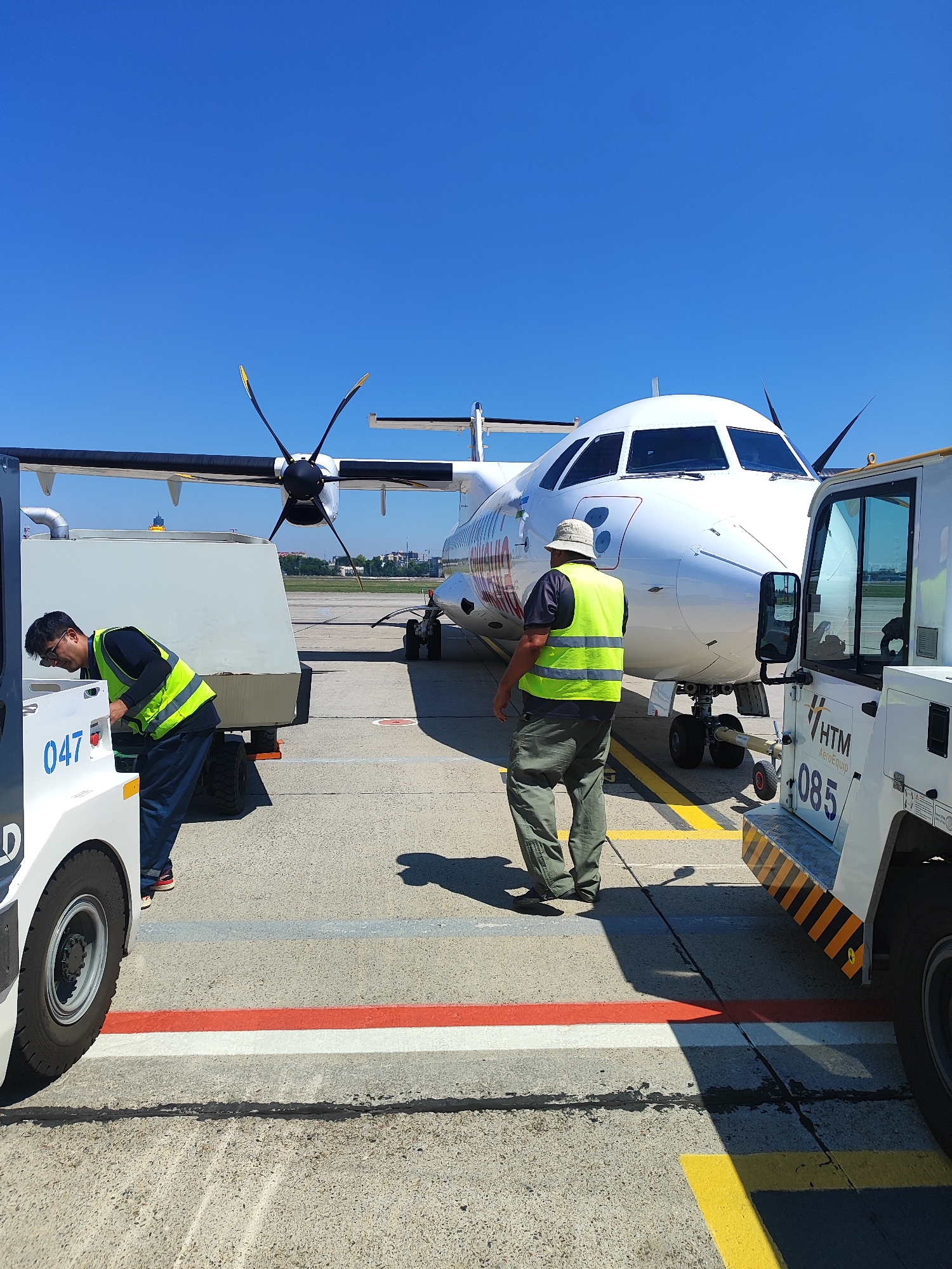 The image shows an airport tarmac on a clear, sunny day. An airplane with two propellers is parked, with its front facing the right side of the frame. Two individuals, wearing high-visibility vests, are working near the plane. One person, on the left, is leaning over a piece of airport equipment that has the number "047" visible. The other person, on the right, is standing and facing away from the camera, looking towards the airplane. Another piece of airport equipment, with "HTM Aeroequip 085" printed on it, is parked to the right of the airplane. The ground is marked with yellow, red, and white lines, typical of airport runway or taxiway markings. The sky is a vivid blue with no clouds.