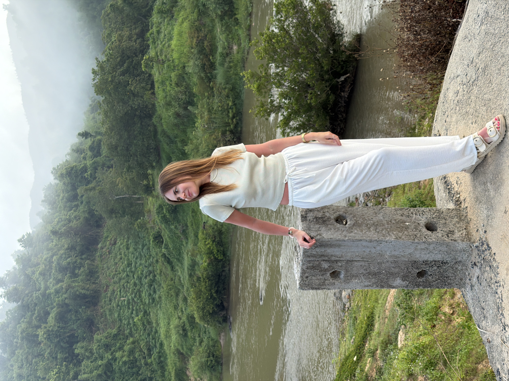 A woman stands on a concrete structure, leaning against a concrete pillar. She is wearing a light-colored crop top and loose white pants, with tan sandals. Her left hand rests on the pillar, and she wears bracelets and a ring. She looks to her right, her head tilted slightly. The background shows a body of water, likely the Nam Khanh Vinh river, with lush green vegetation and misty hills in the distance. The scene appears to be in a natural, somewhat remote setting within Nam Khanh Vinh, Vietnam.
