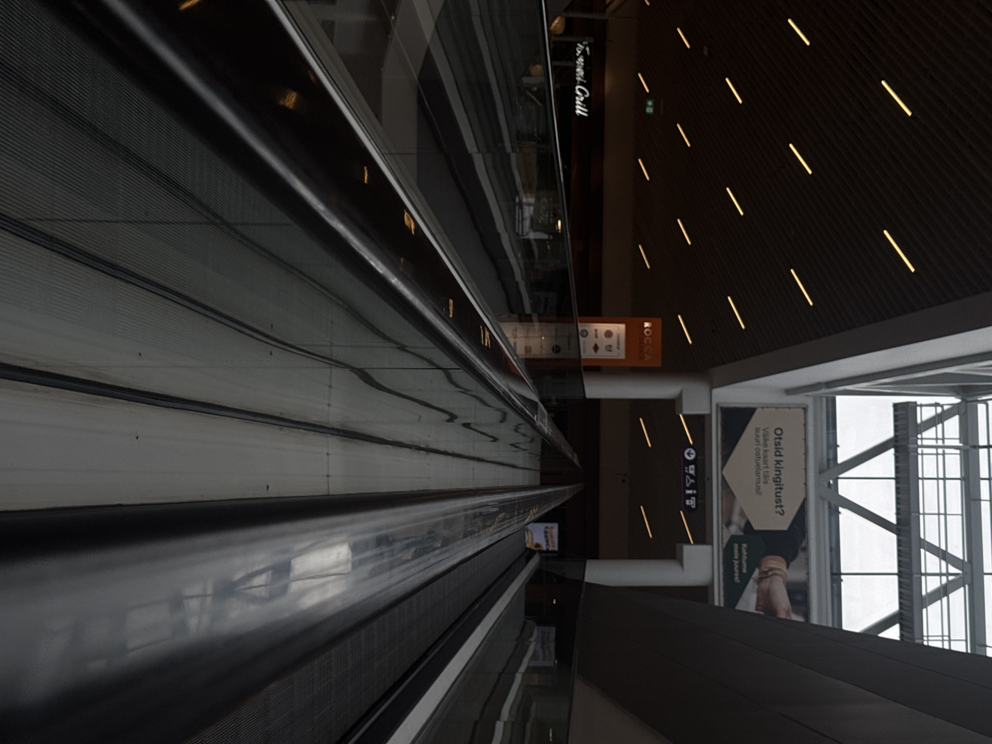 The image shows an interior view of a shopping mall, likely in Tallinn, Estonia, featuring a down-moving escalator. The escalator has a dark grey, metallic handrail and a ribbed grey step surface. The surrounding environment is modern and dimly lit, with rows of yellow strip lights illuminating the dark ceiling. On the left, a glass railing separates the escalator from a lower level, where a sign for "Trattoria Grill" is visible. On the right, another escalator is also visible. In the background, a large advertisement is mounted on a wall, featuring text in Estonian. The overall impression is one of a contemporary, spacious commercial environment.