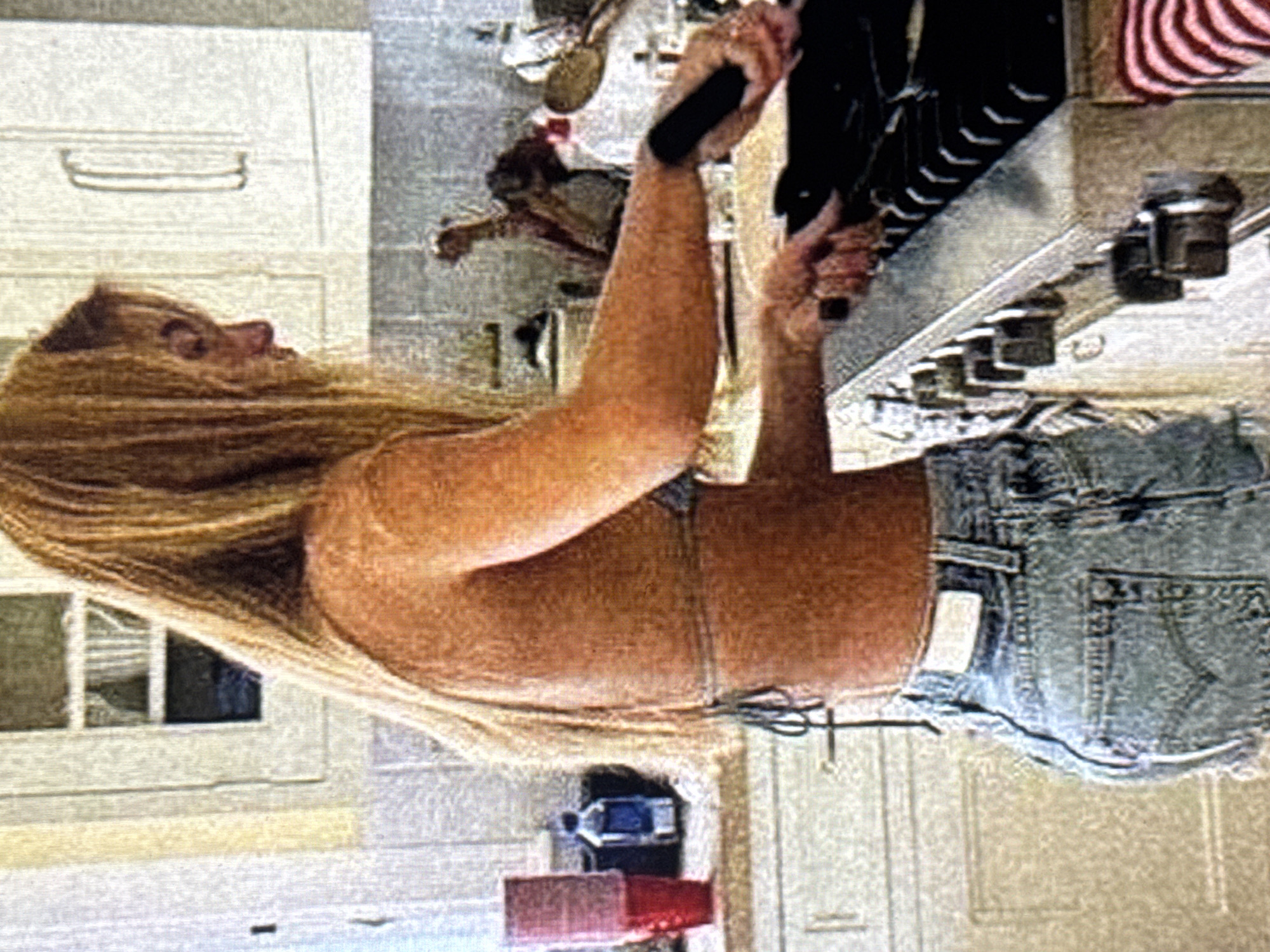 A person with long blonde hair is shown from the side, bending over a counter and holding a black object. The person is wearing denim shorts and a light-colored top. The background appears to be a kitchen with white cabinets and a stainless steel appliance. There is no indication of the time of day or weather. The location cues do not provide specific details to identify the exact setting within Kuta Selatan, Indonesia. No visible text is present in the image.