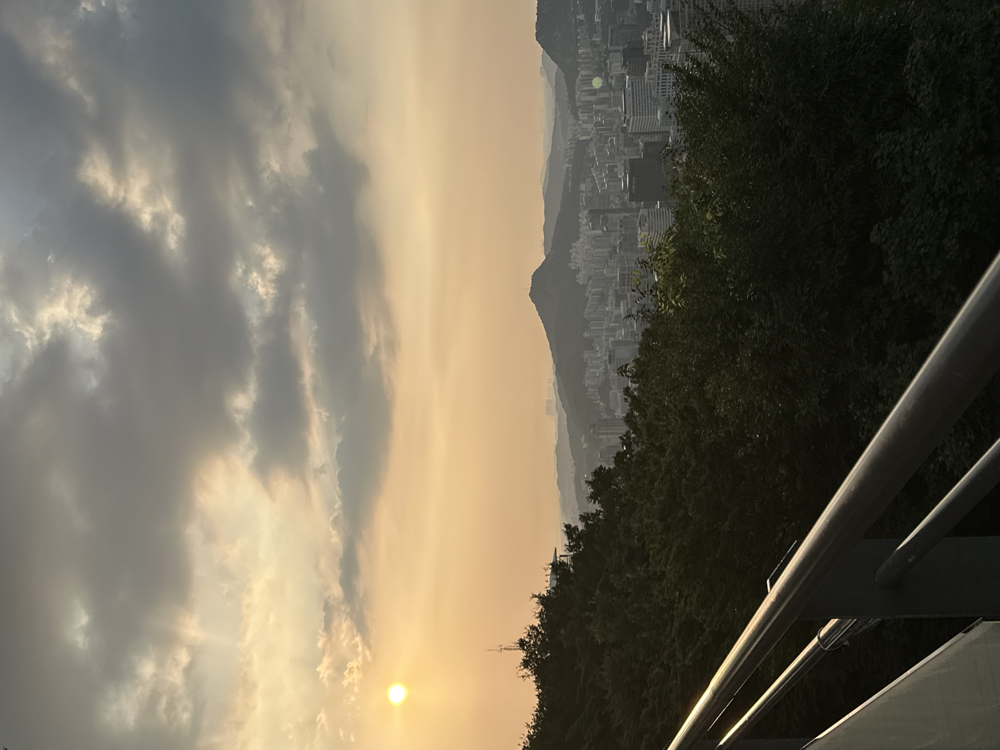 The image captures a panoramic view of Seoul, South Korea, during what appears to be sunset. The sky is filled with dramatic clouds, illuminated by the setting sun in the upper left corner. Rays of light break through the clouds, casting a warm glow over the scene. In the foreground, dark green foliage covers a hillside, with a metal railing running diagonally across the lower portion of the frame. Beyond the trees, a dense cityscape stretches out, filled with buildings of varying heights. In the middle ground, a distinct mountain peak rises above the urban sprawl, a prominent landmark against the hazy horizon. The overall impression is one of a city nestled within a natural landscape, bathed in the ethereal light of dusk.