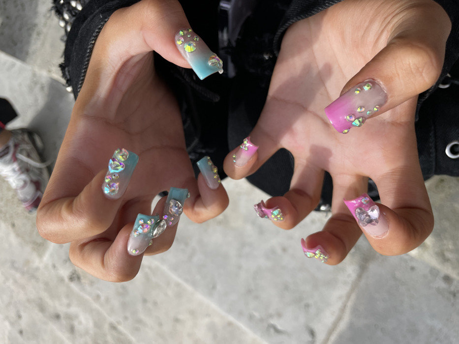 A close-up, high-angle shot captures an individual's hands displaying elaborately decorated fingernails. The specific location within Paris, France, is not discernible from the blurred background, which appears to be a light-colored, textured paved surface, likely stone or concrete, indicating an outdoor setting.
The subject's two hands are positioned in the foreground. The left hand (viewer's left) features long, square-tipped nails with an ombre design transitioning from a clear/white base to a light blue tip. These nails are heavily adorned with multiple iridescent, clear rhinestones of varying sizes. The right hand (viewer's right) also displays long, square-tipped nails with an ombre design, transitioning from a clear/white base to a bright pink tip. These nails are similarly embellished with numerous iridescent, clear rhinestones, including a prominent large one on the thumb. The skin tone of the hands is light to medium brown.
The subject wears dark clothing, possibly a black garment, with visible silver studs or metallic embellishments along the cuff or sleeve. In the upper left background, a blurred white athletic shoe with pink/red accents and white laces is partially visible, resting on the paved surface. The hands are held static, seemingly posed to showcase the intricate nail art.