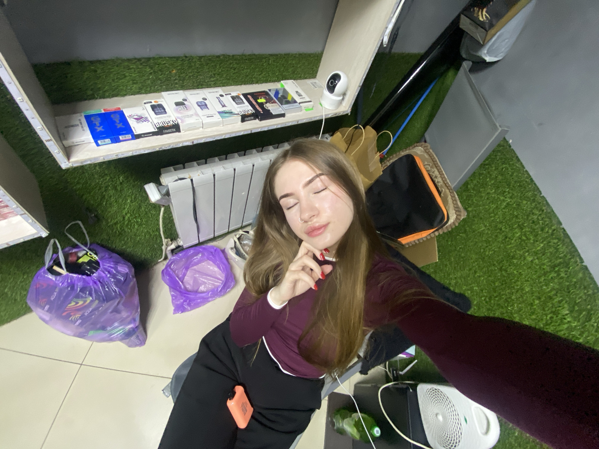 A young woman with long, light brown hair poses for a selfie, looking up with her eyes gently closed and one hand near her chin. She wears a dark red long-sleeved top with white trim and black pants, and has a nose piercing and red nail polish. In her lap, she holds a compact orange electronic device, possibly a vape or charger.
The scene is an indoor space, possibly a retail or storage area, characterized by a mix of white tiled flooring and green artificial turf. A white radiator is visible behind her. On a shelf above her, illuminated by an LED strip light, are several boxed electronic items, including vaping devices and accessories, with visible brand names such as "SMOK," "BRUSKO FAVOSTIX," and "XROS," along with distinctive blue boxes featuring an "X" logo. A white security camera is also positioned on the shelf.
To her left, two purple plastic bags sit on the floor. To her right, near her, are a brown paper bag, a black pouch on a patterned mat, a white circular fan, and a green bottle. The walls are a dark grey, contributing to a utilitarian or modern aesthetic. The lighting suggests an indoor setting, likely with artificial illumination. The overall impression is casual and somewhat cluttered, perhaps reflecting a break or informal moment in a commercial environment.