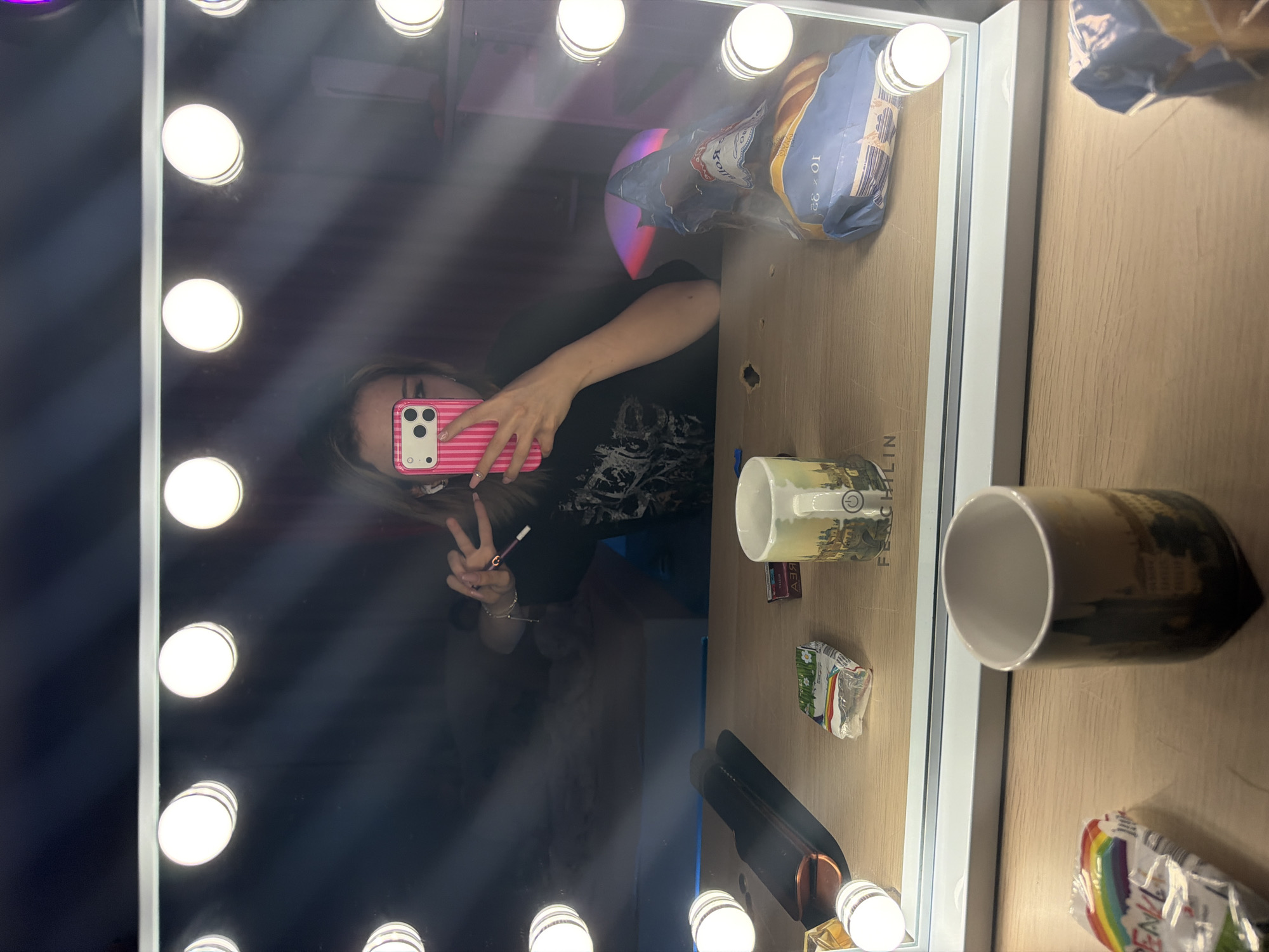 An individual is depicted taking a mirror selfie in an indoor setting, likely a personal vanity or dressing area. The primary subject is the reflection of the individual, appearing to be a young person with light-colored hair, wearing a dark t-shirt with a graphic design. They hold a pink and white striped smartphone (resembling an iPhone) in their right hand, pointed towards the mirror. Their left hand is raised, forming a peace sign, and between the index and middle fingers, a thin, dark cylindrical object, possibly a cigarette or vape pen, is held. White wireless earbuds are visible in both ears.
The mirror is rectangular, framed in white, and features multiple round, illuminated LED bulbs around its perimeter. The vanity tabletop, made of light-colored wood or laminate, holds several items. In the foreground, outside the mirror's reflection, a dark ceramic mug with a cityscape design and a slightly tapered base is visible, alongside a crinkled plastic bag with a rainbow pattern and the word "PENAL."
Within the mirror's reflection on the tabletop, a dark hair styling tool (possibly a flat iron) rests on a black mat. Next to it is a small, white plastic packet with a green floral design. Further right, a small dark box with Cyrillic text "АЭЯ" is present. A second light-colored ceramic mug, also featuring a cityscape and a power symbol on its handle, is visible, with "FERCHILIN" faintly embossed below it. To the far right, a crinkled blue plastic bag, likely containing baked goods, displays text including "Kollo" and "10 x 32."
The background reflected in the mirror appears dimly lit, with a subtle blue glow emanating from an unseen source on the left, and indistinct furniture or shelves on the right.