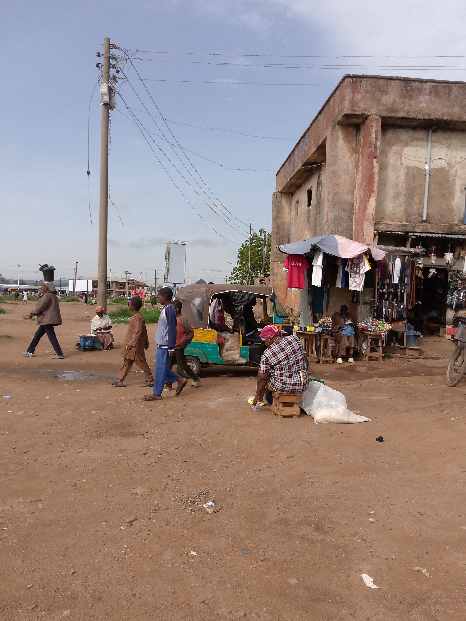 The image depicts an outdoor market scene in Bukuru, Nigeria. In the foreground, several individuals are walking across a dirt road. To the left, a man wearing a brown jacket and dark pants, carrying a black container on his head, walks away from the viewer. To his right, a woman in a red and white checkered shirt and dark pants sits on a wooden stool next to a large white bag. In the midground, a group of children walks past a brown and yellow auto-rickshaw. Behind them, a man in a brown tunic and pants sits at a stall selling goods. Further back, a weathered, light-colored building with a damaged facade stands, with awnings sheltering various items for sale, including clothing and shoes. A utility pole with numerous wires is prominently visible on the left side of the image, extending into the pale sky. The ground is a dry, dusty, reddish-brown dirt surface littered with some debris. The overall atmosphere suggests a bustling, informal marketplace under an overcast sky.