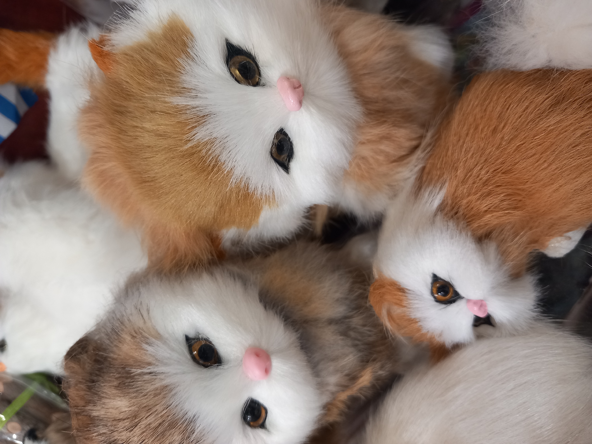 The media file displays a close-up, overhead shot of three stuffed toy cats piled together. The cats are predominantly white with patches of orange fur. They have realistic-looking brown eyes and small, pink noses. Their fur appears soft and fluffy. The composition is tightly cropped, focusing solely on the toys. The background is dark and indistinct, suggesting the toys are likely in a store display or a similar setting. There are no people visible, and no specific activity or event is taking place other than the objects being presented. The lighting is somewhat diffuse, and there are no indicators of time of day or weather conditions. No text is visible in the image.