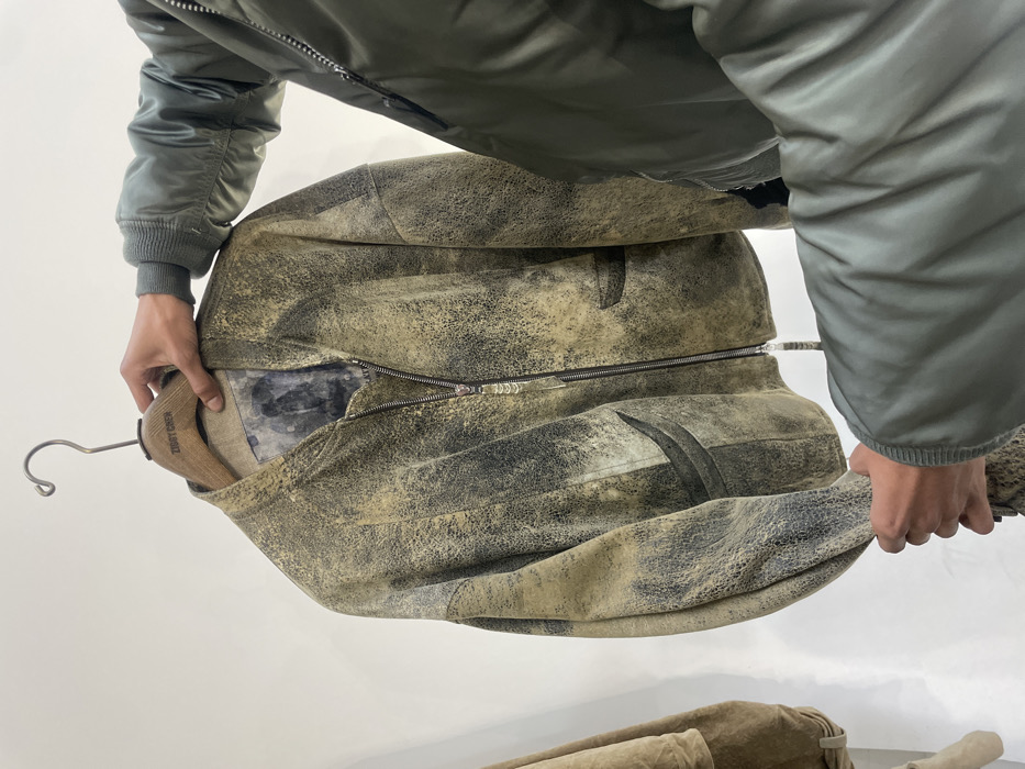 The image displays a person's hands examining a distressed bomber jacket within an indoor setting. The jacket, featuring a mottled pattern of dark green, beige, and black, hangs from a wooden hanger engraved with "ZIGGY CHEN." The person's left hand supports the hanger, while their right hand holds the lower left sleeve of the jacket. They are wearing a light green bomber jacket, visible from the right forearm downwards. A partial view of another light brown garment is visible on the far left. The background consists of plain, light-colored walls, indicating a retail store or showroom. Given the "ZIGGY CHEN" branding, the location is a fashion boutique or showroom in Paris, France.