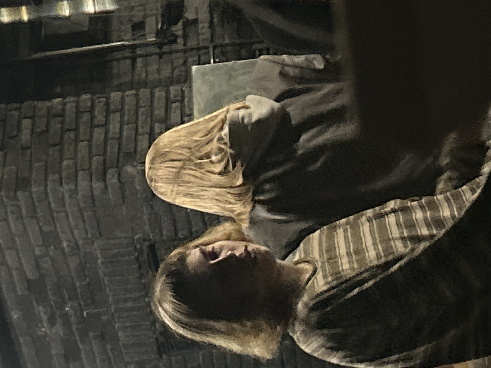 A grainy, slightly out-of-focus image shows two people in what appears to be an indoor setting. The background appears to be a brick wall. A person with light blonde hair is lying down wearing a striped shirt. Above and behind them is another person with similar light blonde hair, wearing a dark shirt and possibly sitting on a square, light-colored object. The location is unidentified.