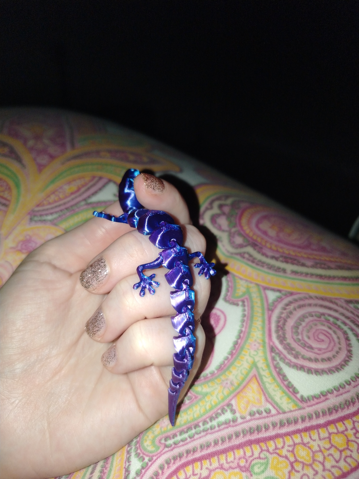 A hand holds a purple and blue articulated lizard. The hand has painted nails with a gold glitter design. The background is partially dark with a paisley patterned fabric visible. The location is not discernible from the image, and therefore cannot be placed in Izhevsk, Russia.