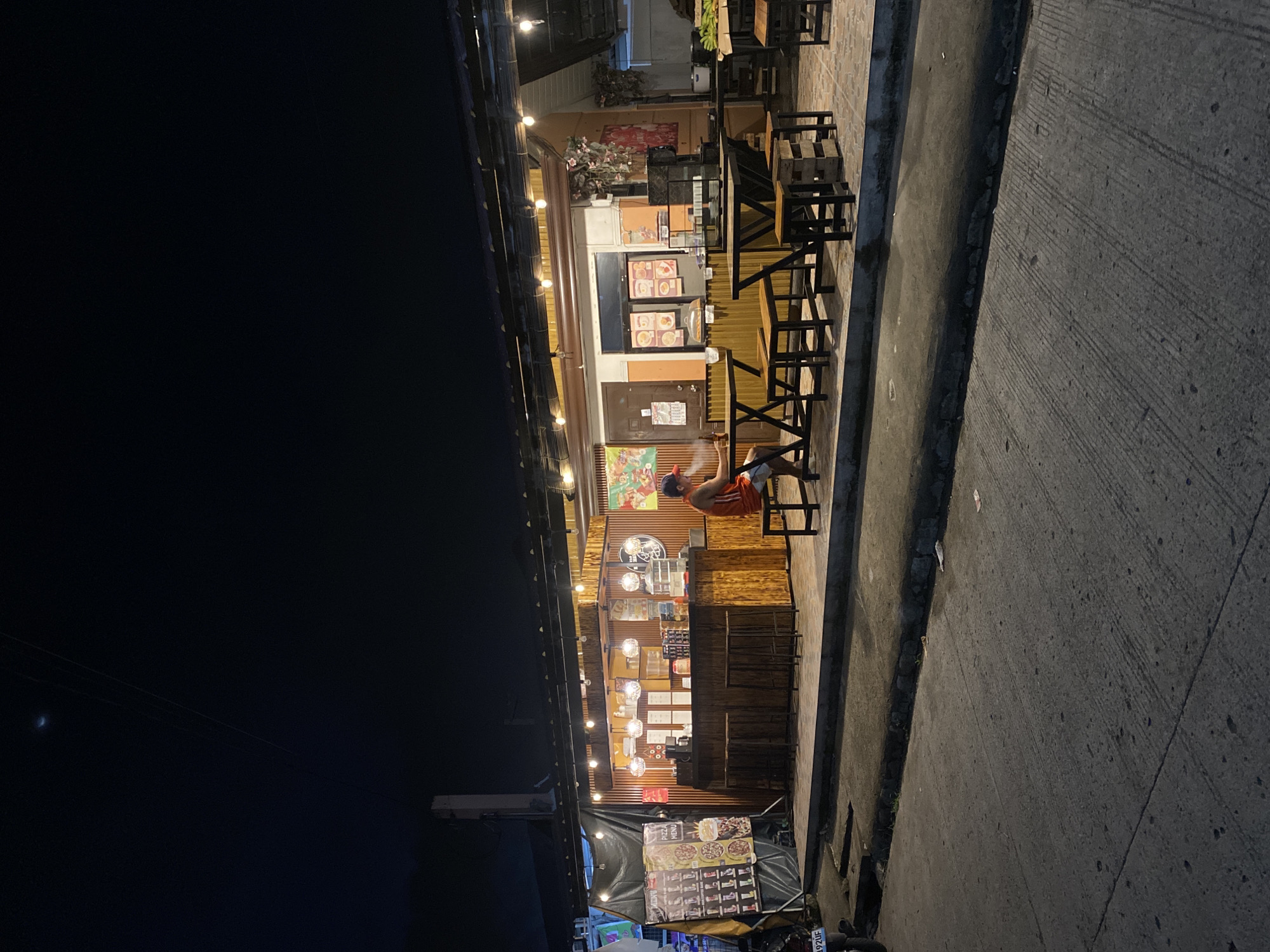 The image shows a small food stall at night, likely located in Davao City, Philippines. The stall has a wooden facade with strings of lights overhead. There are tables and chairs set up outside for customers. A person is sitting at one of the tables, facing away from the camera. The street in front of the stall is paved, and there is a sidewalk. The sky is dark, indicating it is nighttime. There are signs on the stall with menus and what appear to be pictures of food. A motorcycle is parked to the left of the stall.