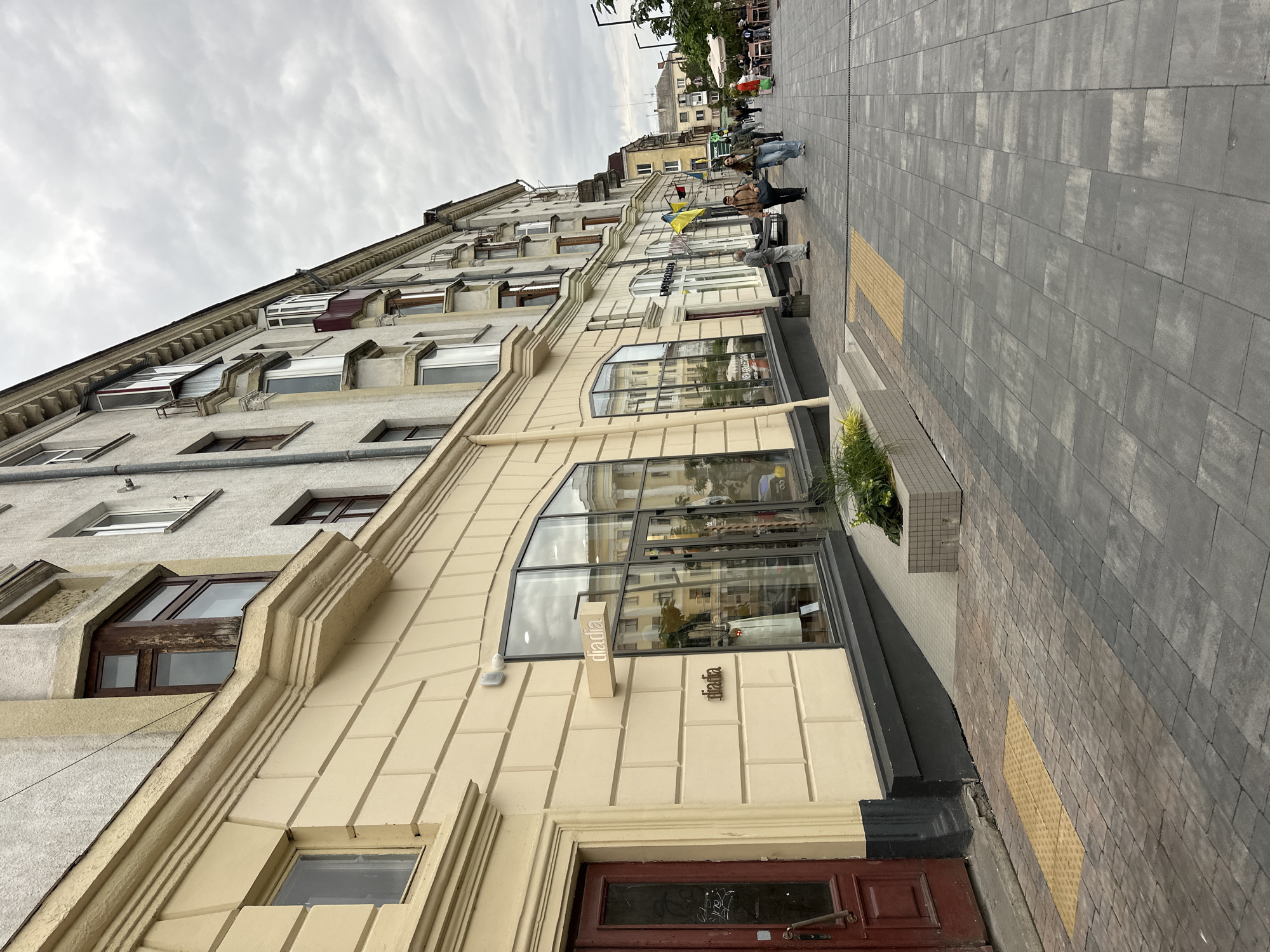 This is a low-angle, eye-level, full shot of a street in Ternopil, Ukraine, on a cloudy day. The primary subject is a cream-colored building with a detailed facade, featuring rectangular stonework and ornate cornices. A prominent storefront with a large glass window is visible, displaying a mannequin dressed in a light green outfit. The store's name, "diadia," is visible in both a raised metallic sign and a smaller illuminated sign above the window. To the left of the storefront is a dark red wooden door. The sidewalk in front of the building is paved with grey interlocking rectangular stones, with a section of textured yellow tactile paving near the building entrance.
Further down the street, the sidewalk is populated by several individuals walking and standing. Two young adults, a man and a woman, are walking towards the viewer, while an older man stands near the storefront. In the background, more people are seen sitting at outdoor tables, suggesting a cafe or restaurant area. Two Ukrainian flags, one larger and one smaller, are prominently displayed on poles attached to the building further down the street. Other buildings line the street, varying in architectural style and color, with a mix of windows and balconies. Trees and awnings are also visible in the background. The overall atmosphere is that of an urban street scene with commercial activity.