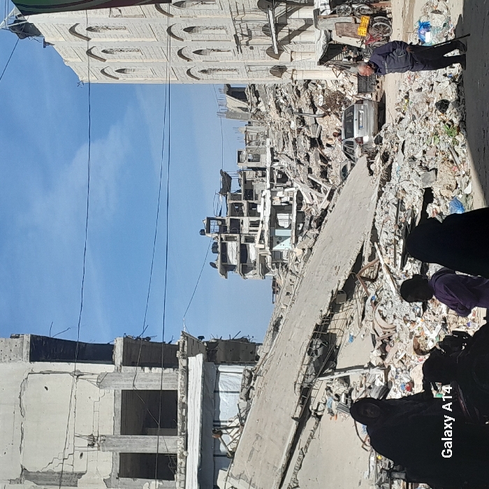 The image portrays a scene of widespread urban destruction in Khan Yunis, Palestinian Territory, under a clear blue sky. The foreground and mid-ground are dominated by extensive piles of concrete rubble, broken slabs, and twisted rebar from collapsed buildings.
On the left side of the foreground, three individuals are partially visible. One person, dressed in dark clothing, looks towards the viewer. A child or young adult in a light purple top stands with arms crossed, facing right. Another individual in dark clothing is partially obscured. On the right, an older man with short grey hair, wearing a dark jacket and pants, stands using a walking stick, looking towards the left.
The mid-ground features a light-colored sedan partially visible behind a significant mound of debris. Multiple multi-story buildings in the background exhibit severe damage, appearing as skeletal concrete structures with missing walls, exposed interiors, and collapsed sections. One structure on the far left shows extensive facade damage and internal exposure. On the far right, a multi-story building with an ornate light-colored facade, featuring arched windows and columns, appears relatively intact on its visible side, adjacent to further rubble. A bicycle and a handcart with yellow crates are visible near this building. Several thin utility lines crisscross the sky overhead.