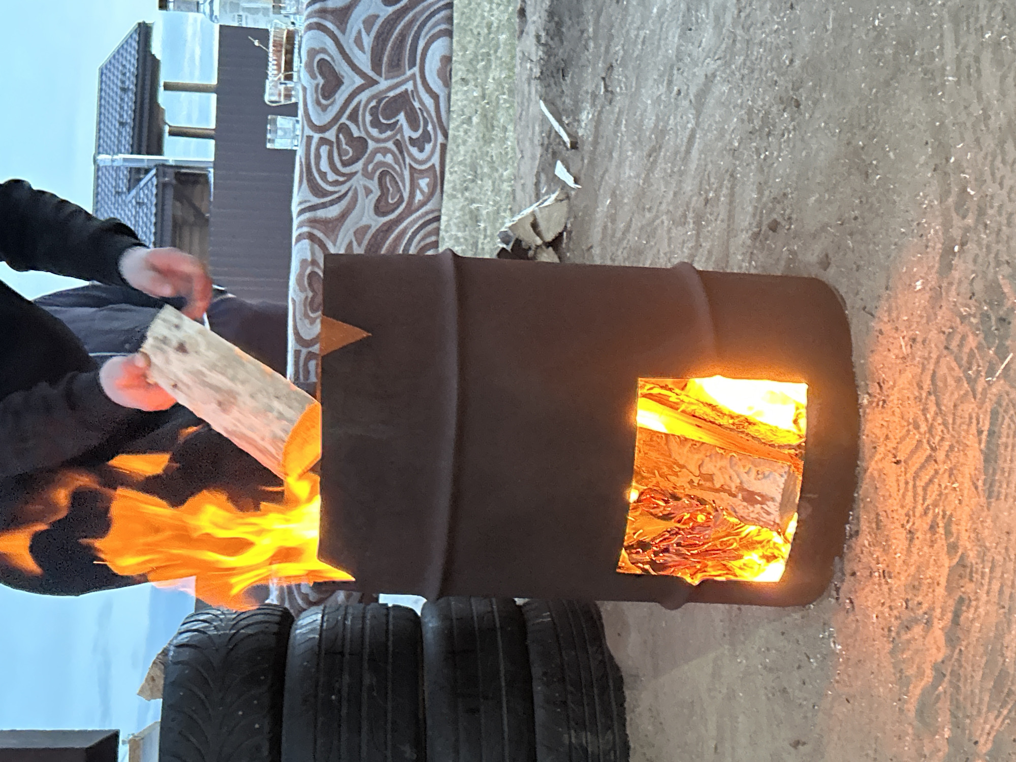 An outdoor scene in Noviy, Russia, depicts a person feeding a fire contained within a modified metal barrel at dusk. The barrel, dark brown and standing on dirt ground, features a rectangular cut-out at its lower front, revealing bright orange and yellow flames consuming wood inside. Larger flames emanate from the barrel's open top. To the left, a stack of three dark vehicle tires is visible. A partially visible individual, wearing a dark long-sleeved garment, holds a light-colored piece of wood, positioning it above the barrel's opening, suggesting they are adding it to the fire. Behind the barrel and person, a table draped with a brown and white heart-patterned tablecloth displays a clear glass bottle, several clear glasses, and a dish with dark contents. In the background, beyond a dark fence, a building with a dark tiled roof is visible against a light blue sky. Scattered pieces of firewood lie on the ground near the barrel.