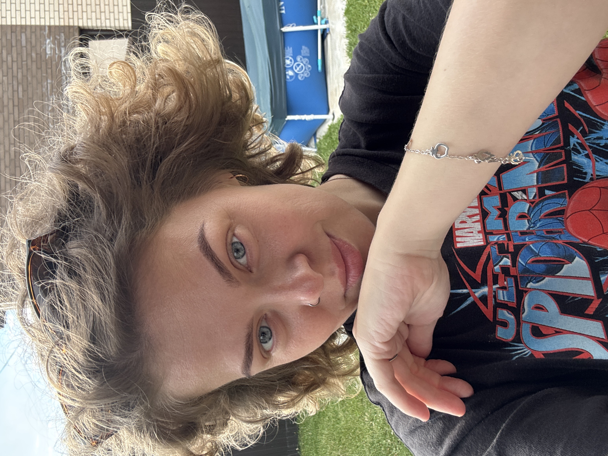 A woman with curly blonde hair and blue eyes is shown from the chest up, looking directly at the camera. She is wearing a black t-shirt with a graphic print. Her right hand is resting on her chin, and she is wearing a silver bracelet on her left wrist. The background shows a green lawn, a blue inflatable pool, and a brick building. The image appears to be taken outdoors on a sunny day. The location of Дёмино, Russia is not discernible from the image.
