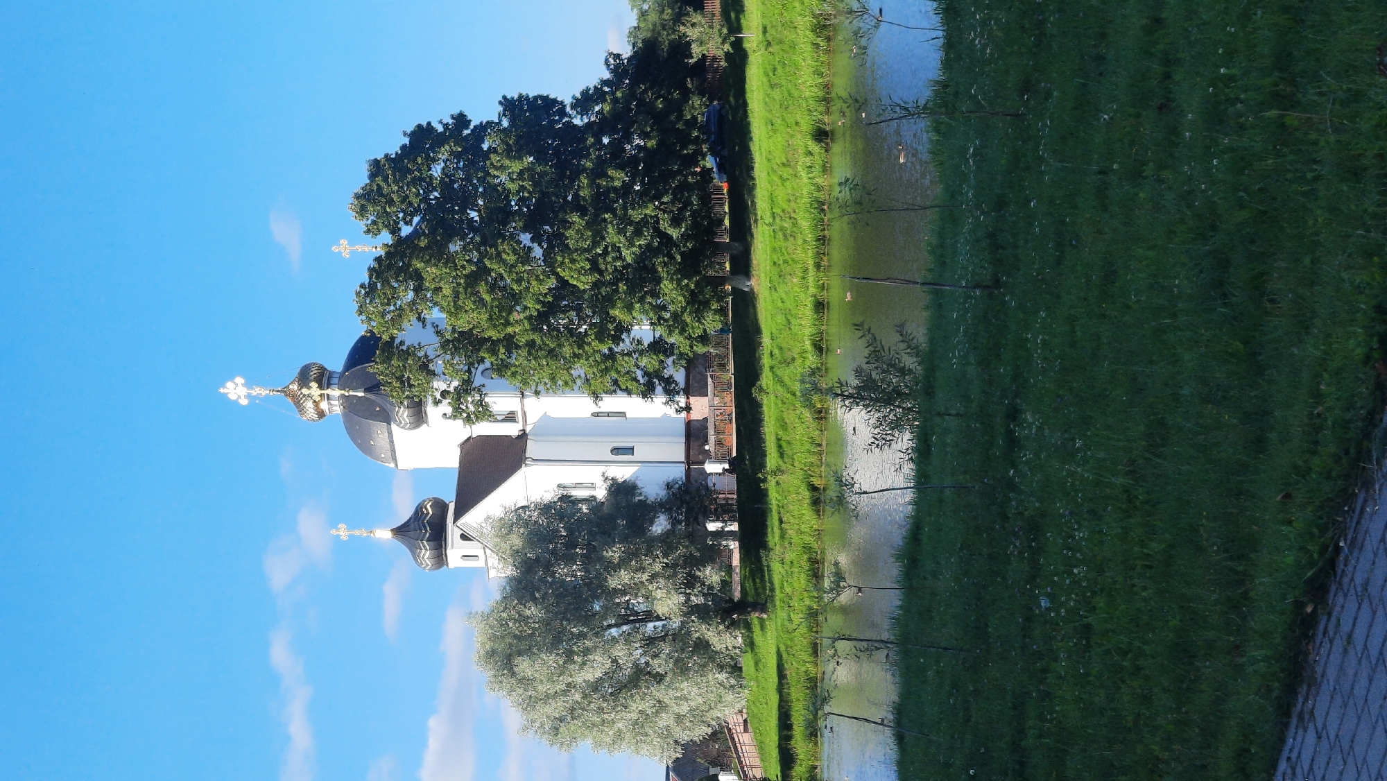 The image shows an Orthodox church with multiple golden domes and crosses, set against a bright blue sky with scattered white clouds. The church is white with dark roofs and domes. It is surrounded by lush green trees, with a prominent silver-leafed tree on the left and a large, dark green tree on the right. In the foreground, there is a body of water, possibly a river or lake, with some ducks swimming. Beyond the water, a grassy embankment slopes up towards the church. The embankment has some small saplings planted in rows. The overall scene depicts a serene, well-maintained landscape with the church as the central focal point. This is the Holy Protection Orthodox Church, located in the city of Smarhon, Belarus.
