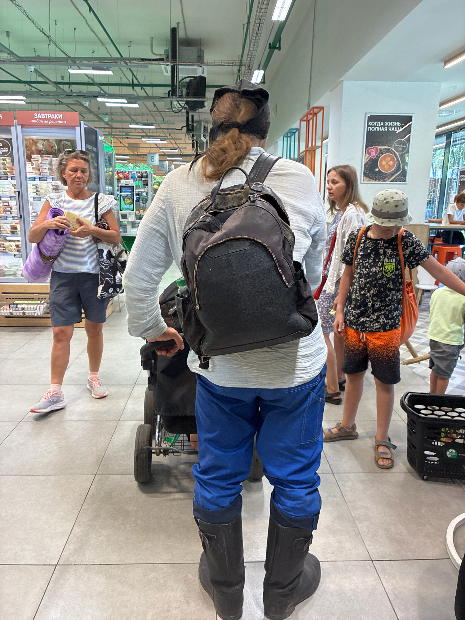 This is a medium shot, eye-level, full-body photograph taken indoors, likely in a grocery store or supermarket. The foreground is dominated by the lower half of a person wearing blue trousers and black boots, pushing a stroller or shopping cart. The stroller has two visible wheels and a black basket.
Behind this person, to the left, a woman with graying blonde hair pulled back is holding a yellow item and a purple rolled-up mat, and a tote bag with a black and white cat pattern. She is wearing a striped t-shirt and grey shorts. She appears to be looking down at the items in her hands.
Further back, to the right of the central figure, a young boy wearing a bucket hat and a dark t-shirt with orange shorts is visible. He has a bright orange bag slung over his shoulder. A younger child in a light green t-shirt and grey shorts is standing to his right.
In the background, more people are visible, and shelves stocked with products can be seen. The environment suggests a retail setting with fluorescent lighting overhead and a tiled floor. There is a poster on a white pillar that reads "КОГДА ЖИЗНЬ - ПОЛНАЯ ЧАША" (When life is a full cup) with an image of a cup of coffee. The overall scene depicts a group of people shopping or moving through a public commercial space.
The specific location in Sochi, Russia, is indicated by the text on the poster.