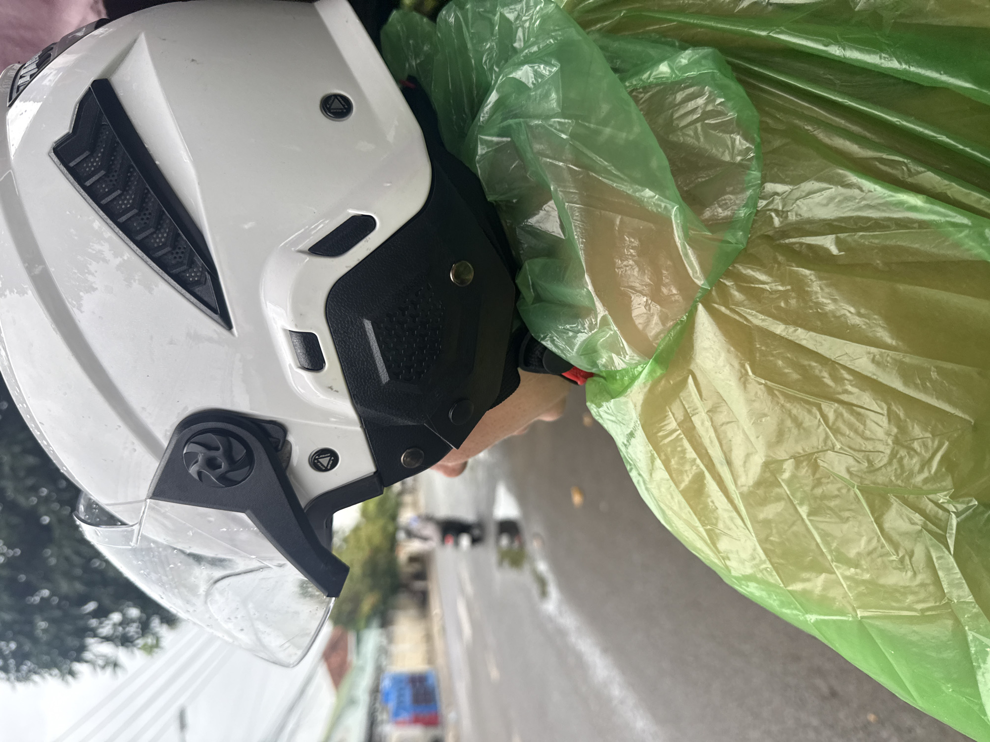 A person wearing a white motorcycle helmet and a translucent green raincoat is seen from behind, looking towards a wet street in Nha Trang, Vietnam. The street is partially covered in puddles, indicating recent rain. In the background, a person on a motorbike is visible further down the road, also riding in the wet conditions. Buildings and trees line the street. The overall impression is that of riding a motorcycle in rainy weather.