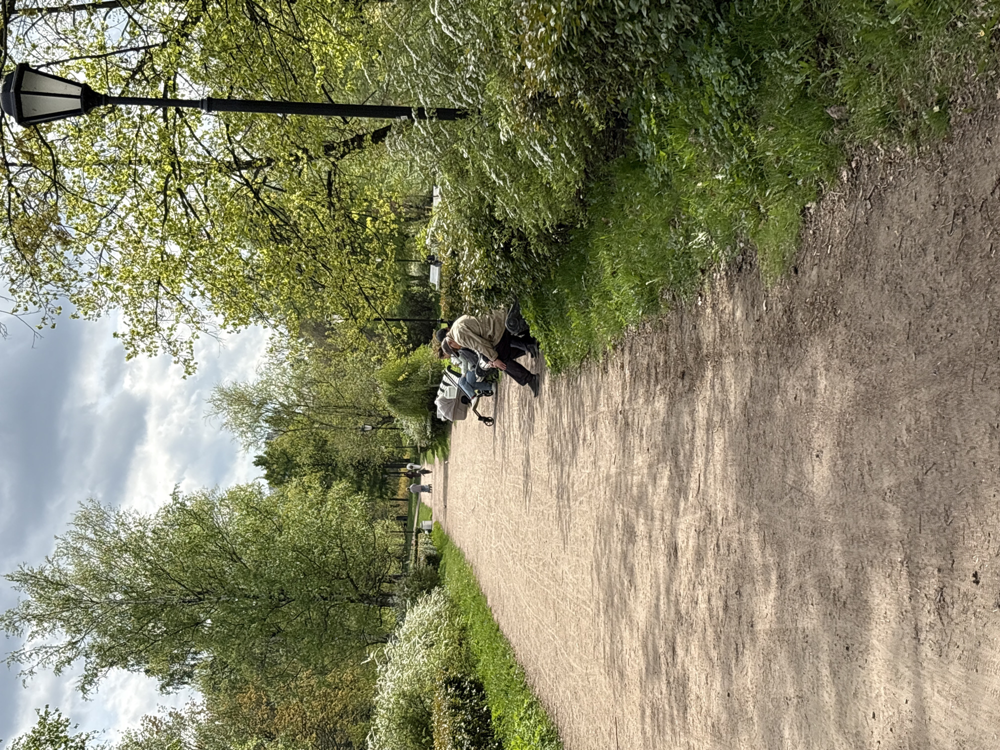 The image depicts a park scene, likely located in Saint Petersburg, Russia, as indicated by the distinctive style of the lamppost and the presence of foliage typical of the region. A dirt path stretches into the distance, lined on either side by trees and greenery.
Two individuals are sitting on a bench to the right of the path. One person, dressed in dark pants and a light-colored jacket, is bent over, possibly attending to something on the ground. Another person sits next to them.
In the background, further down the path, a figure can be seen walking. Benches are visible along the path. The weather appears overcast, with a mix of cloudy skies. The scene suggests a peaceful park setting with people engaging in leisurely activities.