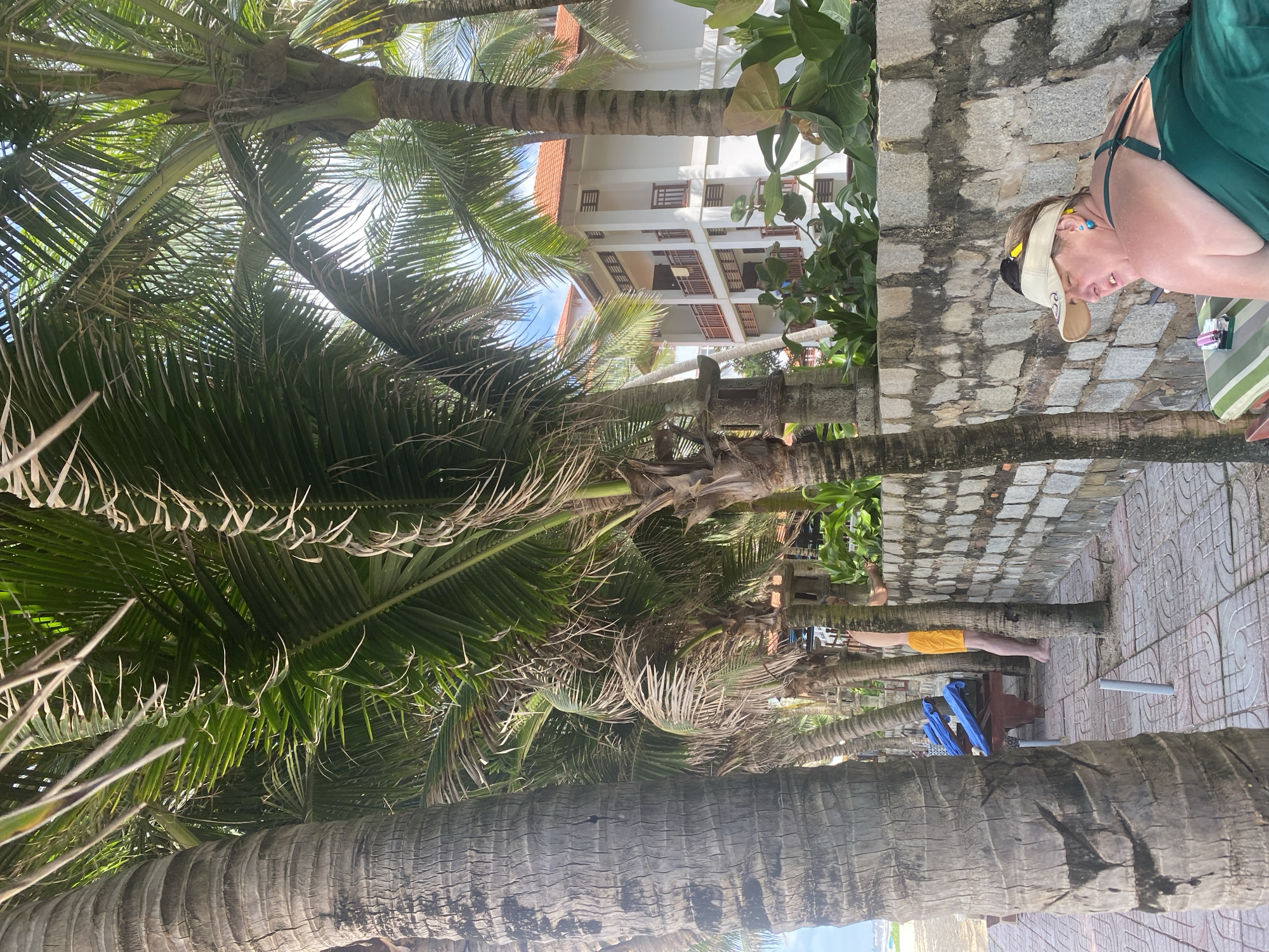 The image shows a tropical scene with prominent palm trees in the foreground. In the background, a multi-story white building with balconies and terracotta-tiled roofs is visible, suggesting a hotel or resort. The setting appears to be outdoors, possibly a beach or a walkway leading to one, given the paving stones and lush greenery.
A woman wearing a green swimsuit and a visor is partially visible on the right side of the image, reclining. Her eyes are closed, and her head is tilted back, suggesting relaxation or rest. Another person, wearing yellow shorts, is seen standing near a palm tree in the mid-ground. Two blue chairs are placed near a wooden bench.
The lighting suggests it is daytime, with sunlight filtering through the palm fronds. The weather appears to be warm and sunny, typical of a tropical climate. The presence of palm trees and the architectural style of the building strongly point to a location in Phu Thuy Ward, Vietnam. No visible text is present in the image.