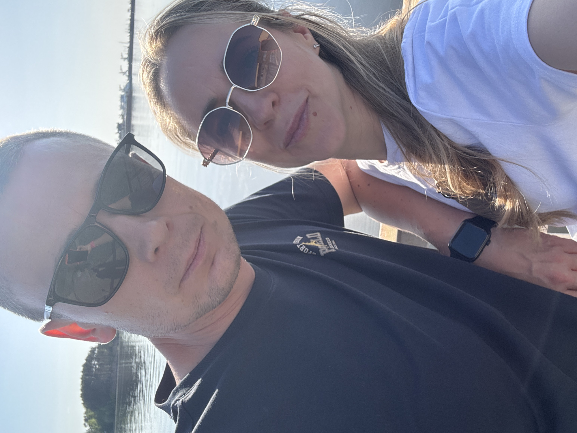 A selfie shot angled downward, featuring a man and a woman. The man, on the left, has short gray hair, light skin, and wears black sunglasses and a black t-shirt. The woman, on the right, has long blonde hair, light skin, and wears round sunglasses and a white t-shirt. She also has a black smartwatch on her left wrist. They are close together, possibly on a pier or elevated platform. The background includes water, greenery, and what appears to be the skyline of Chaikovsky, Russia.