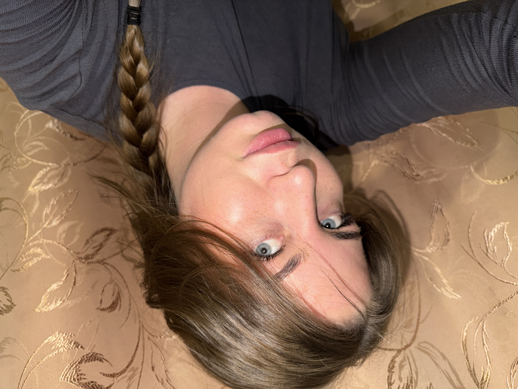 The image is an upside-down selfie of a Caucasian woman with light skin, light blue eyes, and light brown hair. Her hair is partially braided and partially loose, falling across her face. She is wearing a dark gray, long-sleeved shirt. The background is a tan-colored patterned fabric, possibly a bedspread or upholstery. The location is not discernible from the image, and there is no information to suggest it is in Chaikovsky, Russia. There are no discernible actions or interactions within the image, it is a portrait of the subject's face.