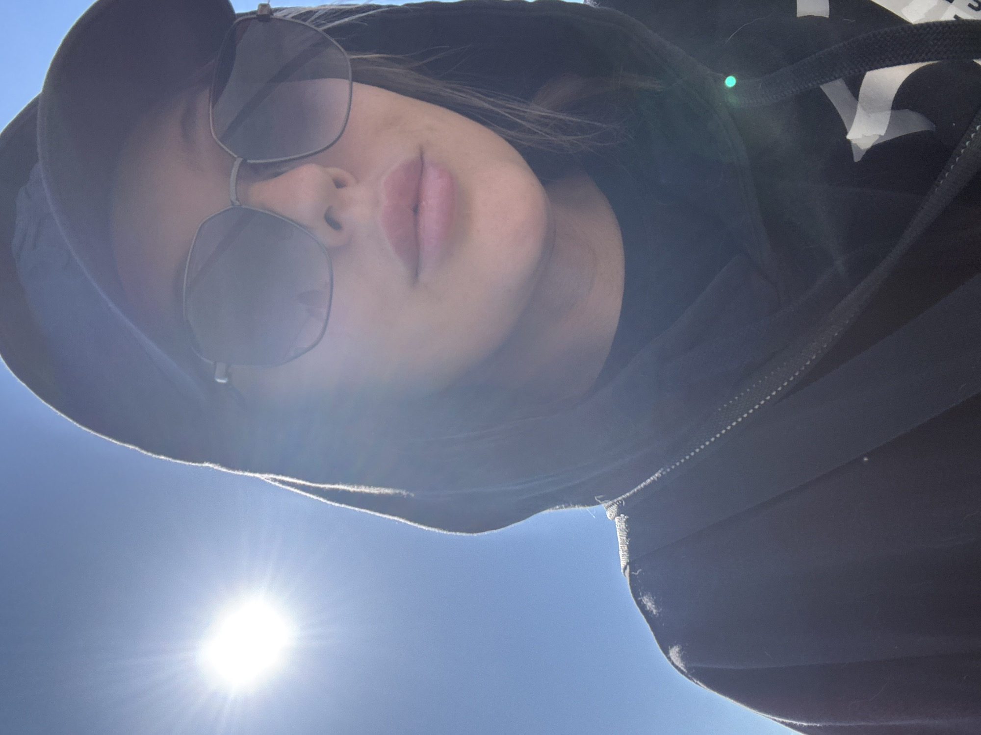 A person wearing a black hooded sweatshirt and sunglasses looks down at the camera. The sun shines brightly in the sky behind them. The location cannot be determined from this image.