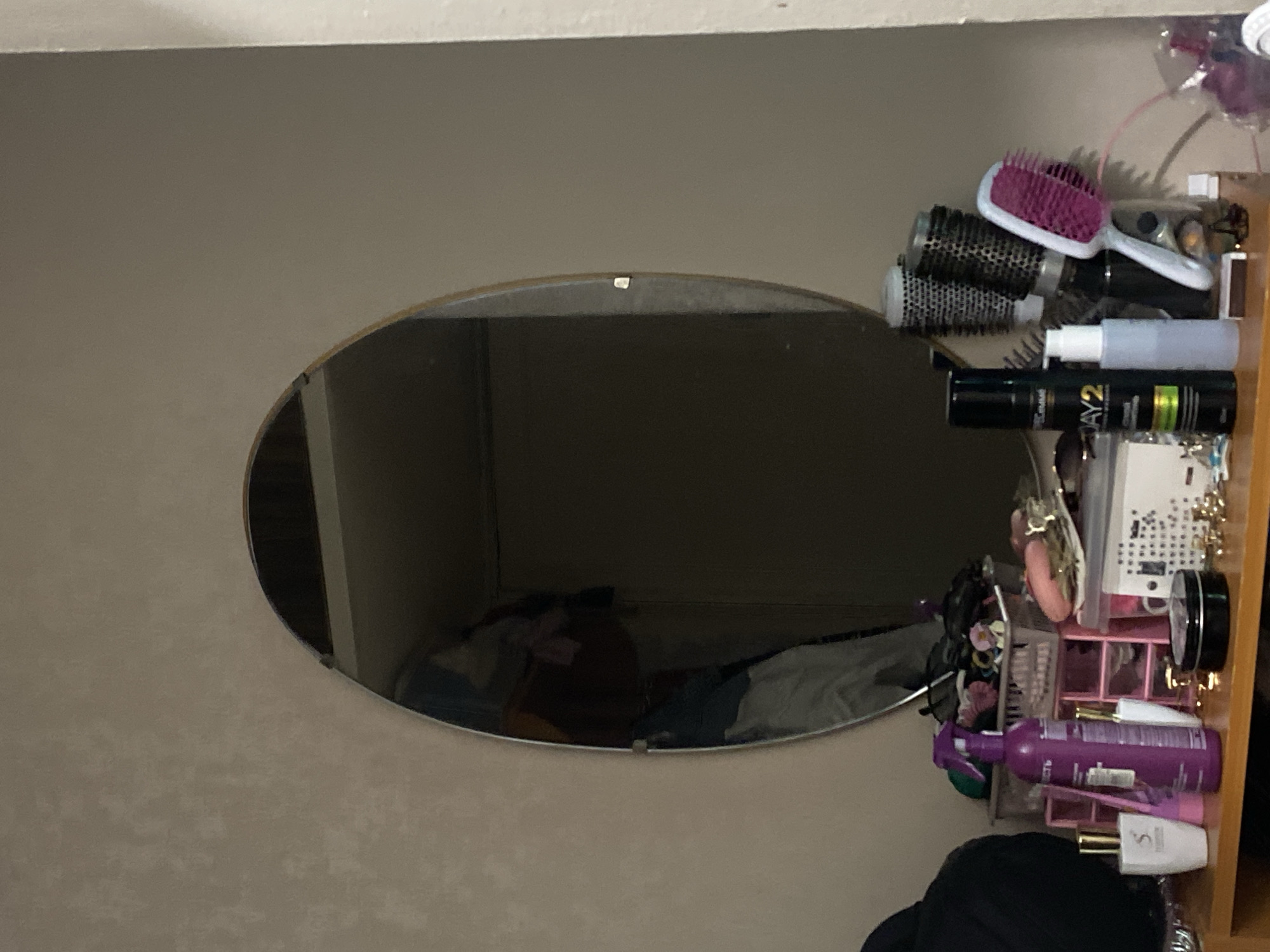 The image depicts a vanity table with various cosmetic and hair styling products. An oval mirror hangs above the vanity. On the vanity, there are several bottles, brushes, and containers. A person's reflection is visible in the mirror, wearing a maroon shirt and gray pants. The background appears to be a wall. The scene suggests a personal grooming area. The image was taken in Lomonosov, Russia.