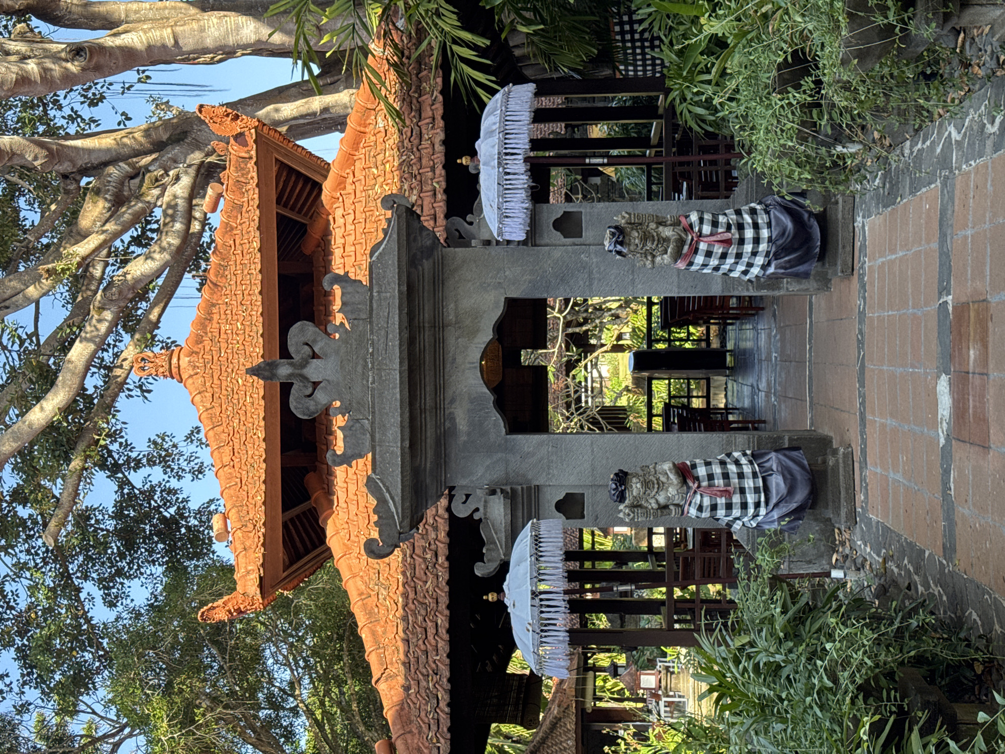 A Balinese-style temple gate stands in Kuta Selatan, Indonesia, with ornate, terra-cotta tiled roofs and stone statues flanking the entrance. The statues are of mythical figures, adorned in traditional black and white checkered sarongs and red sashes. White ceremonial umbrellas with tasseled fringes stand next to them. Beyond the gate, a covered outdoor dining area is visible with tables and chairs. Lush green vegetation, including potted plants and creeping vines, surrounds the pathway leading to the gate. A large, mature tree with exposed roots and hanging aerial roots looms in the background, its branches reaching towards the clear blue sky. The sun casts shadows, suggesting it is daytime.