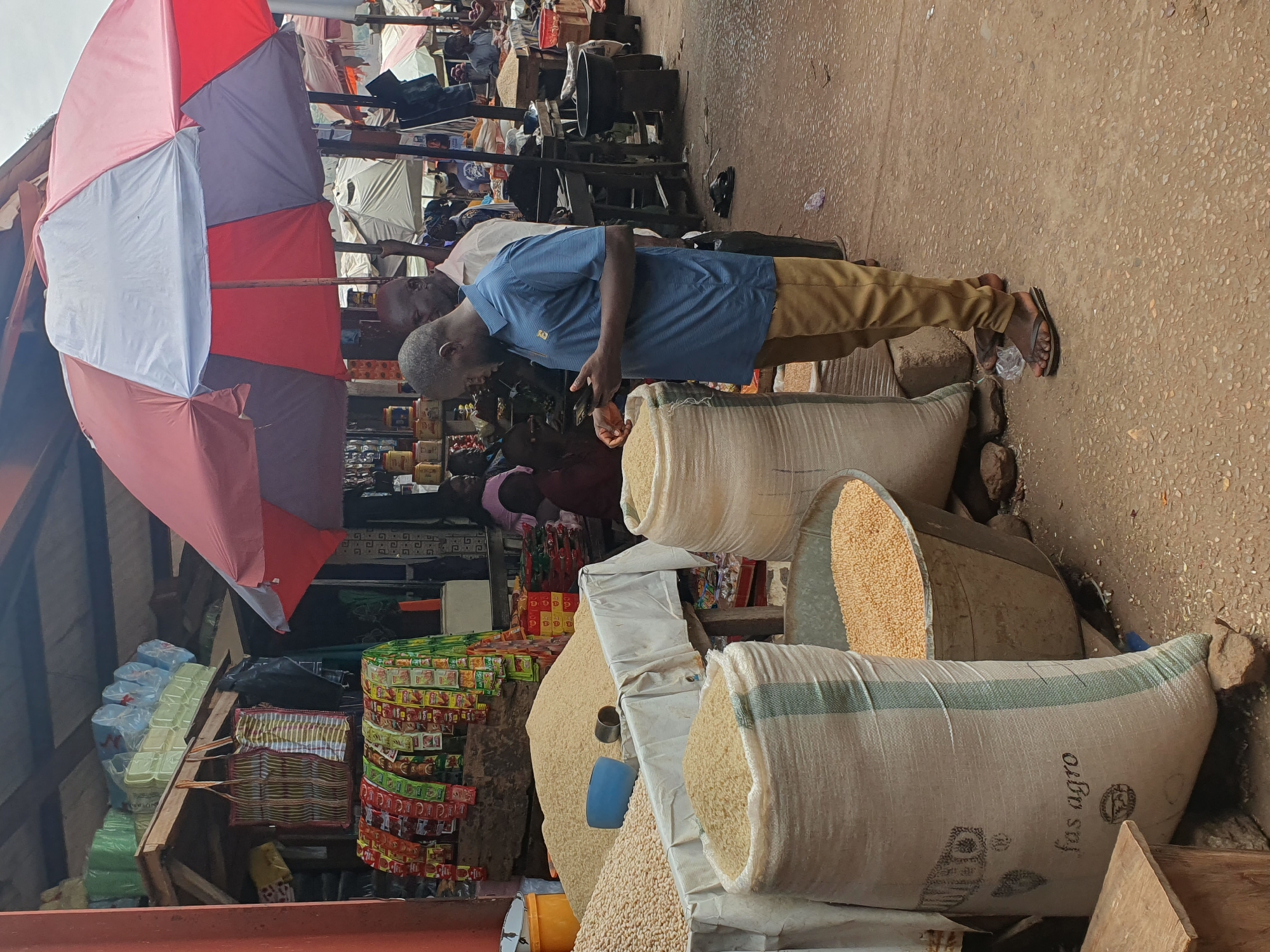The image shows a bustling open-air market in Lokoja, Nigeria. An elderly man in a blue short-sleeved shirt and khaki pants is examining grains from a large sack. The sacks are filled with grains, likely rice or corn. A red and white umbrella provides shade to vendors selling goods stacked on makeshift shelves. Items for sale include food packets, seasonings, and water bottles. Other people are moving about in the background. The ground is a light brown color, likely a combination of dirt and concrete.