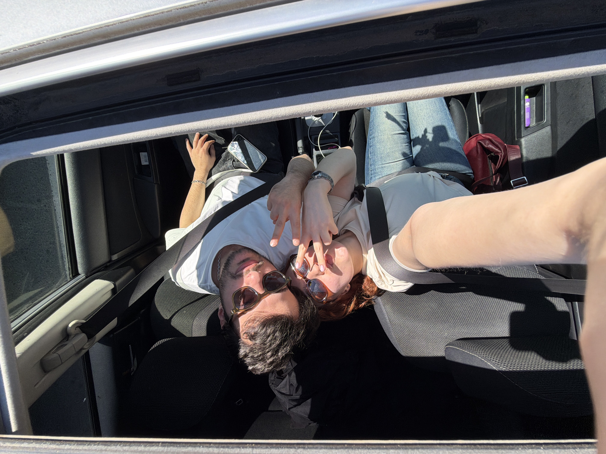 Two individuals are positioned within the front seats of a car, visible through an open sunroof. The man on the left wears a white t-shirt and sunglasses. The woman, wearing a wristwatch, mirrors the man's attire with a light-colored shirt and similar sunglasses. Their hands are raised, partially obscuring their faces. The car's interior features black seats and a visible seatbelt across each person's chest. The vehicle is assumed to be in transit in Tbilisi, Georgia, though definitive landmarks are absent in the picture.
