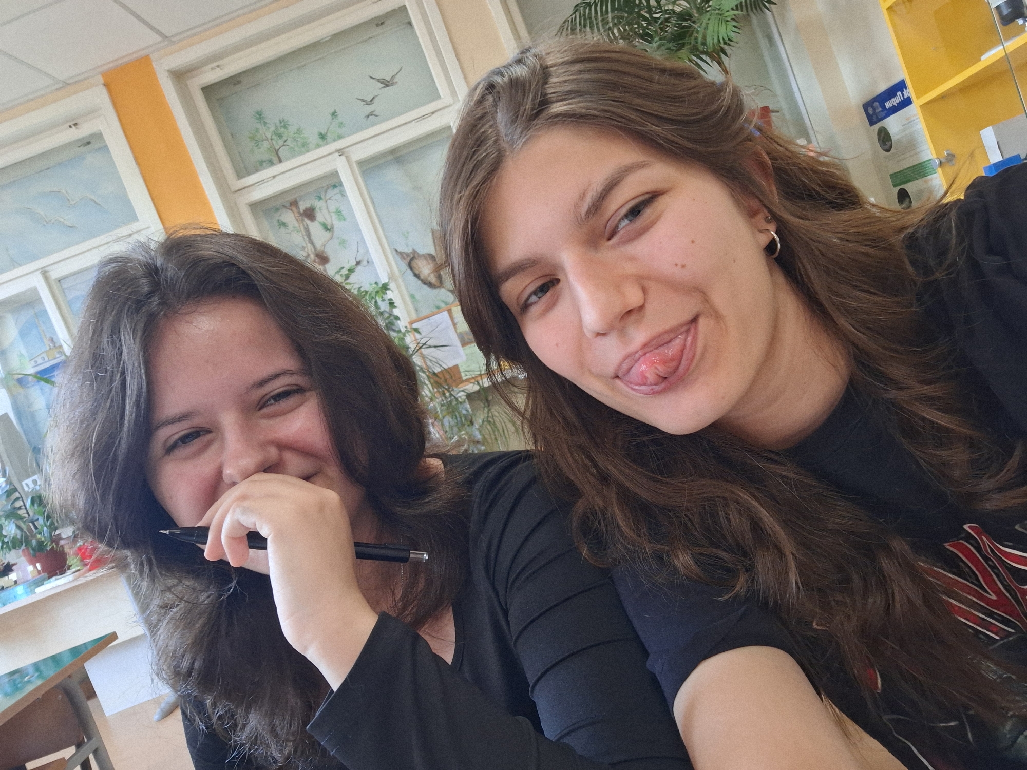 Two young women are posing for a selfie indoors. The woman on the right is sticking her tongue out. The woman on the left is smiling and holding a pen near her mouth. Both women have long brown hair and are wearing dark clothing. Behind them is a window with artwork depicting nature scenes and birds. A yellow cabinet is visible on the right side of the image. Based on the visible writing in Cyrillic on the cabinet, and confirmed by other visual data, it is presumed that the location is in Bulgaria, likely in the city of Panagyurishte, in a classroom or similar educational environment.