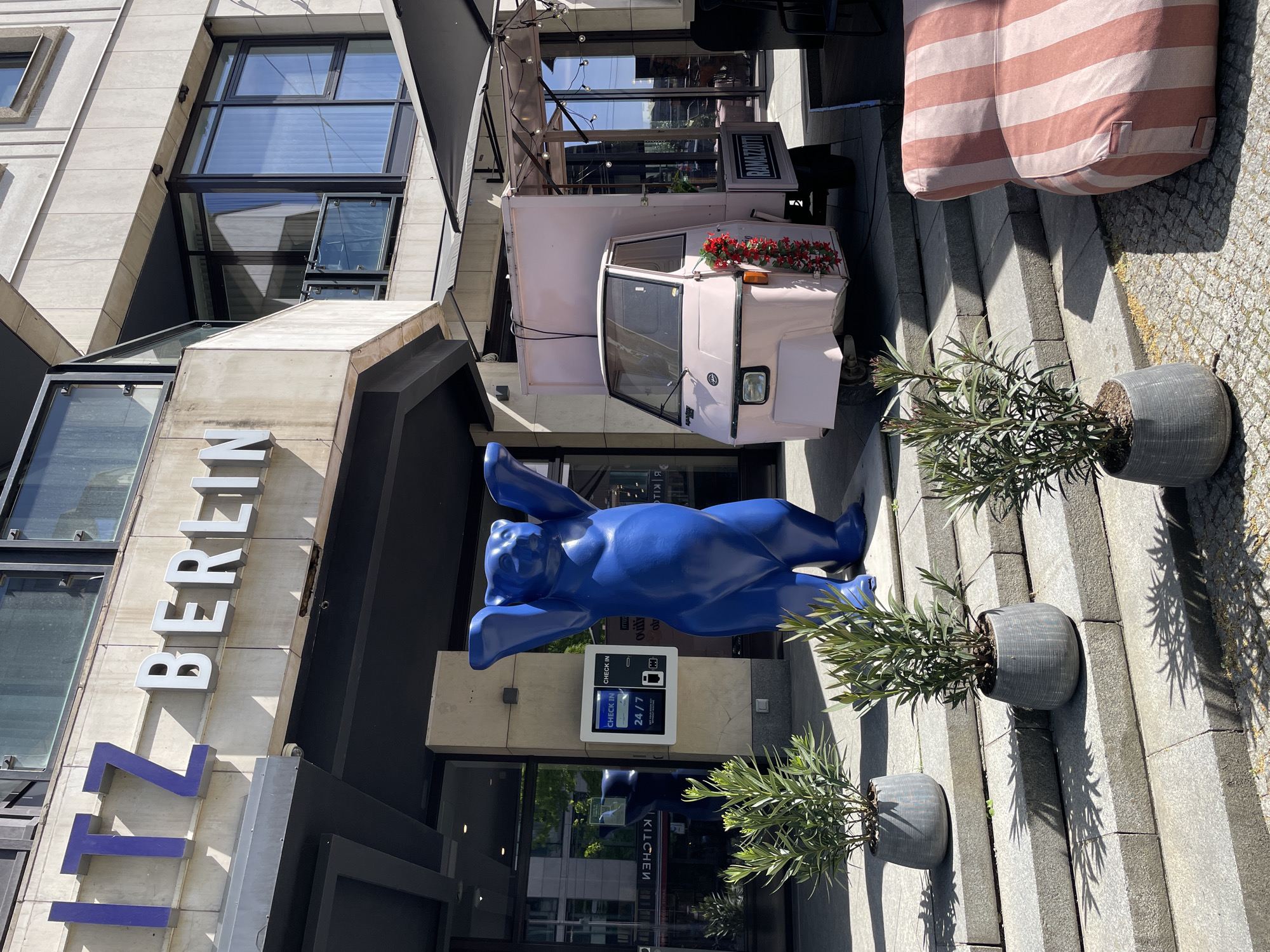 The image shows the 112 Hotel Berlin, Germany. A blue bear statue stands in front of the entrance, flanked by potted plants. A pink Piaggio Ape with red flowers sits on the sidewalk next to striped patio furniture. The building has modern architecture with large windows. A self check-in kiosk is visible at the entrance.