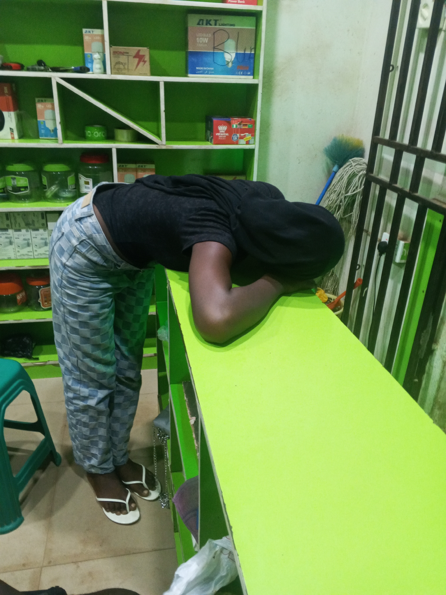 The image shows a person, whose face is not visible, leaning over a bright lime green counter. They are wearing a black hooded sweatshirt and checkered grey and blue pants. White flip-flops are visible on their feet. The person appears to be resting or possibly asleep.
Behind the counter are shelves stocked with various items, including what appear to be light bulbs in boxes, jars, and other small products. Some of the boxes have text on them. One box on the top shelf displays "AKT LIGHTING" and "LED BULB 10W", and "MADE IN CHINA" in English and Arabic. Another box on the same shelf has a red lightning bolt symbol. On a lower shelf, there is a red and white box with "WONDERFUL" printed on it.
To the right of the shelves, a mop with a blue handle and a dirty string head rests against a white wall. A dark metal grate structure is also visible to the right of the mop. The overall setting appears to be a small shop or store. The lighting suggests it could be daytime, but the interior lighting is also evident. There are no obvious cues about the weather. The scene evokes a sense of fatigue or a quiet moment within the shop.