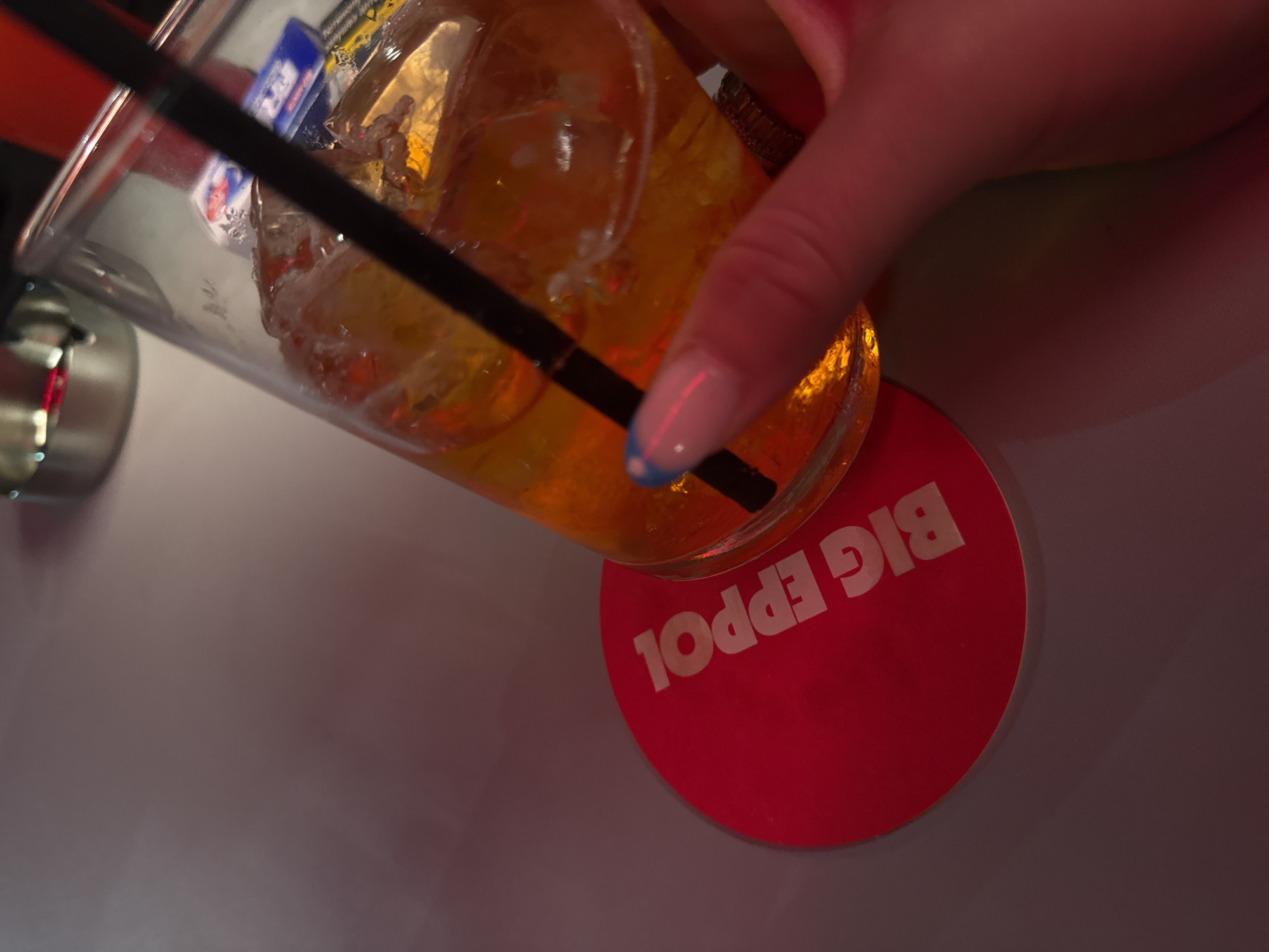 A tilted glass of amber liquid filled with ice and a black straw sits on a red coaster with white lettering reading "BIG EPPOL" in Milan, Italy. A hand with manicured nails grips the glass. Part of a metallic object is visible on the left side of the frame, near the glass. The surface below the coaster is light gray.