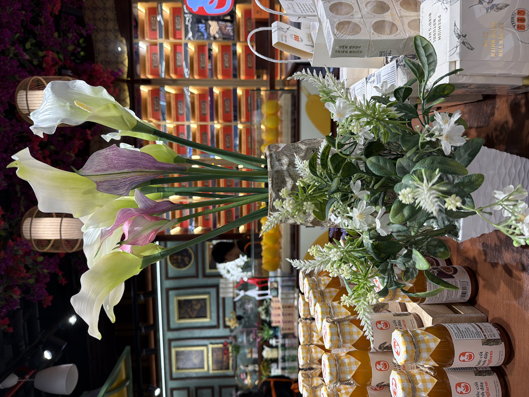 This image depicts a close-up view of a retail display, likely in a shop in Da Nang, Vietnam. The foreground is dominated by rows of small glass bottles filled with an orange liquid, possibly juice or concentrate, with checkered yellow and white lids. Next to them, a white vase holds an arrangement of artificial flowers, primarily white calla lilies with hints of pink, and smaller white wildflowers and green foliage. In the background, shelves are filled with stacks of boxes, many with red and orange accents, suggesting packaged goods. A person's silhouette is visible behind a glass counter in the mid-ground, facing away from the camera. The environment appears to be indoors, with decorative lighting fixtures and wall art visible in the upper and mid-ground, contributing to a market or boutique atmosphere.