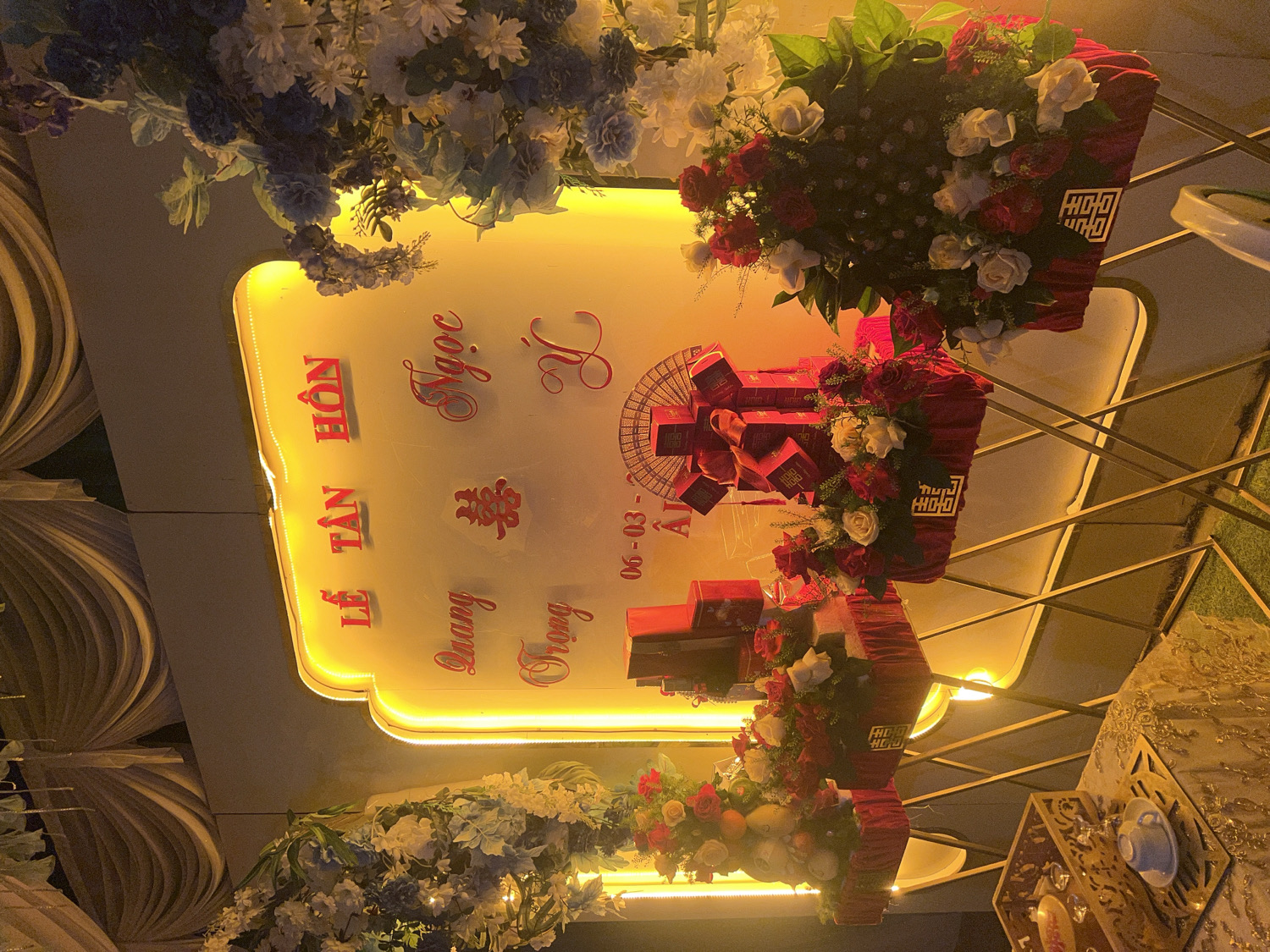 This image captures a vibrant and elegantly decorated scene for a wedding or engagement ceremony, set indoors, likely in Bình An, Vietnam. The ambiance is warm and festive, characterized by abundant lighting and floral arrangements.
The central feature is a large, illuminated sign with a glowing yellow border and light background. Prominently displayed in red, stylized Vietnamese script are the words "LỄ TÂN HÔN," which translates to "Wedding Ceremony." Below this, the names "Ngọc" and "Quang Trọng" are visible, presumably the names of the couple. A traditional Chinese "Double Happiness" character (囍) is also featured on the sign. Part of a date, "06-03-AI," suggesting March 6th, is also present.
Lush floral decorations are integrated throughout the display. On the top left, clusters of white, blue, and purple flowers cascade downwards. On the right side of the sign, two large, opulent arrangements of red and white roses, intertwined with dark green foliage, are positioned on tiered stands. Beneath these floral displays are several red, rectangular gift boxes, some adorned with gold accents and the "Double Happiness" symbol, likely representing traditional offerings or wedding favors.
To the far left, the background features decorative panels with a wavy, layered texture. On the far right, a small table covered with an intricate gold and white patterned tablecloth holds a delicate white teacup with a blue rim, resting on a decorative golden stand. The overall lighting, originating from the glowing sign and other hidden sources, casts a soft, celebratory glow over the entire setup, indicating an evening event. The combination of traditional symbols, Vietnamese text, and elaborate decor creates an atmosphere of joy and solemnity appropriate for a significant matrimonial occasion.