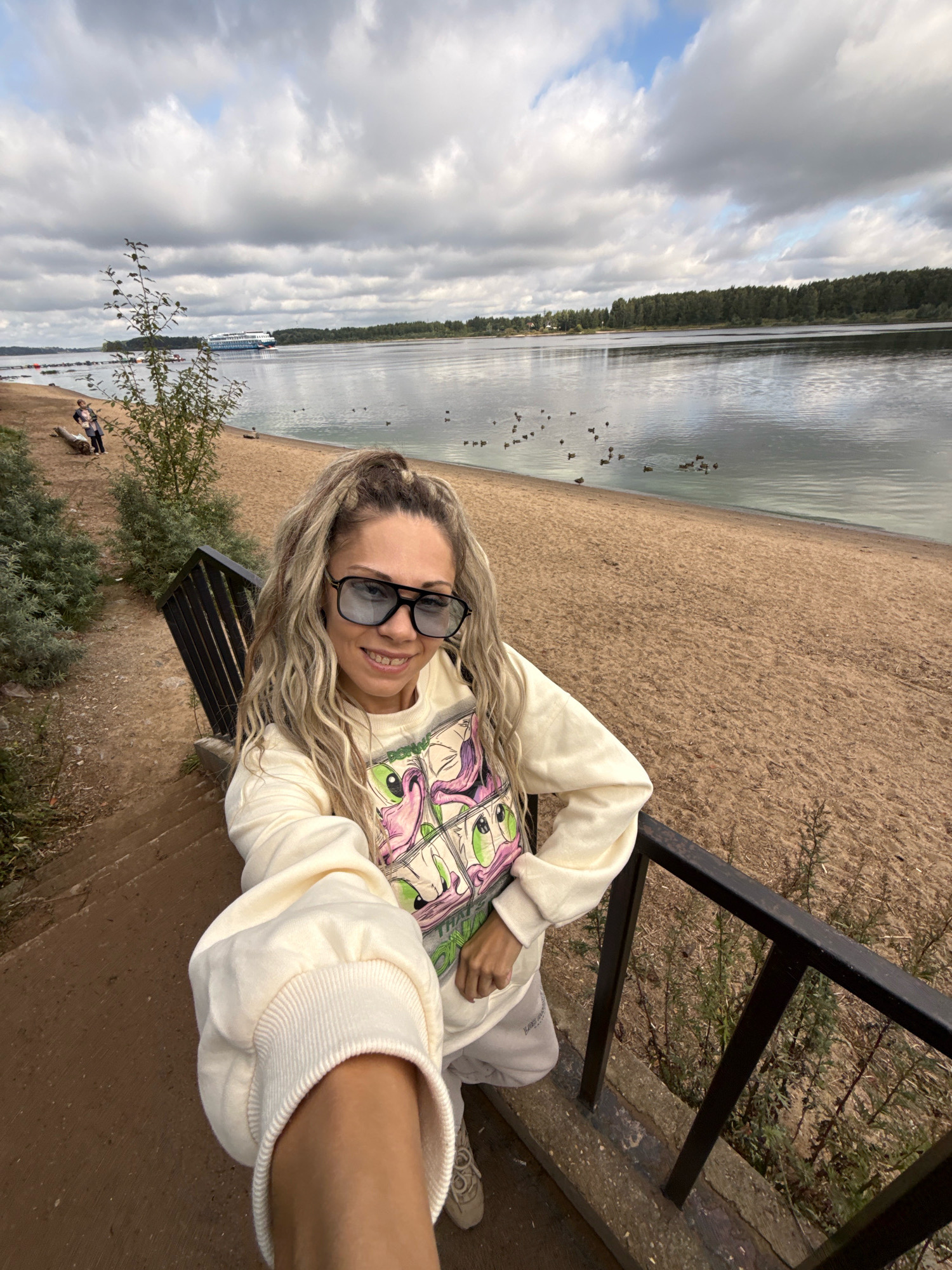 In Rusanovo, Russia, on a cloudy day, a woman with blonde, curly hair pulled into a high ponytail, wearing oversized sunglasses, a light-colored sweatshirt with a cartoon print, and light-colored sweatpants, smiles at the camera, taking a selfie. She is standing on a set of outdoor stairs with a black metal railing, which leads down to a sandy beach. The beach borders a large body of water, possibly a river or lake, where several ducks are swimming. In the distance, a large white and blue cruise ship is visible on the water, and a tree line forms the horizon. The overall environment suggests a recreational area with a natural landscape.