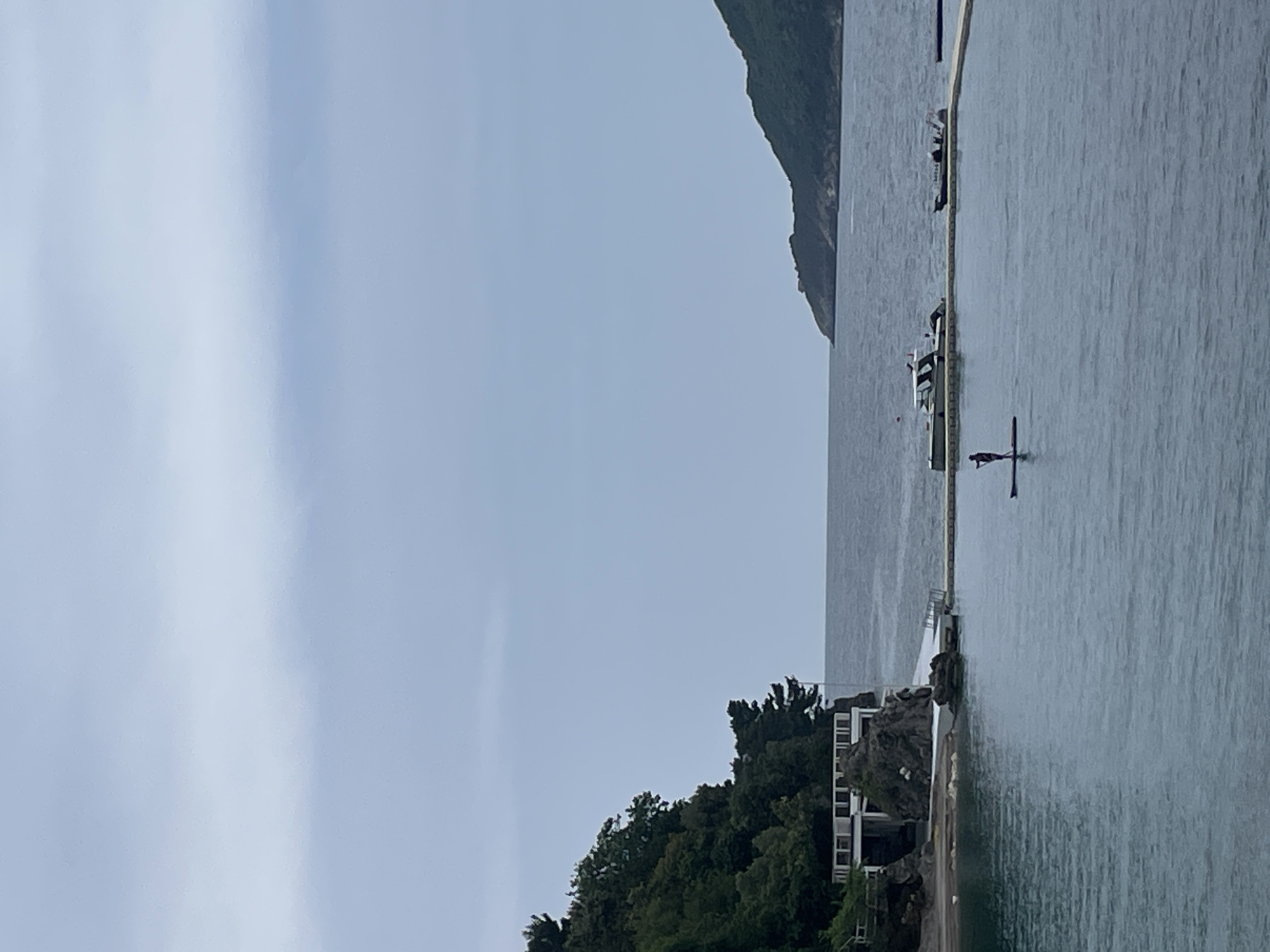 The image shows a coastline in Budva, Montenegro. On the right is the Adriatic Sea, and a person stands on a paddleboard in the water. Further in the distance, boats are docked near a boom. The shoreline is marked by a building with trees, with an adjacent rocky promontory. In the distance is a mountain range. The sky is overcast.
