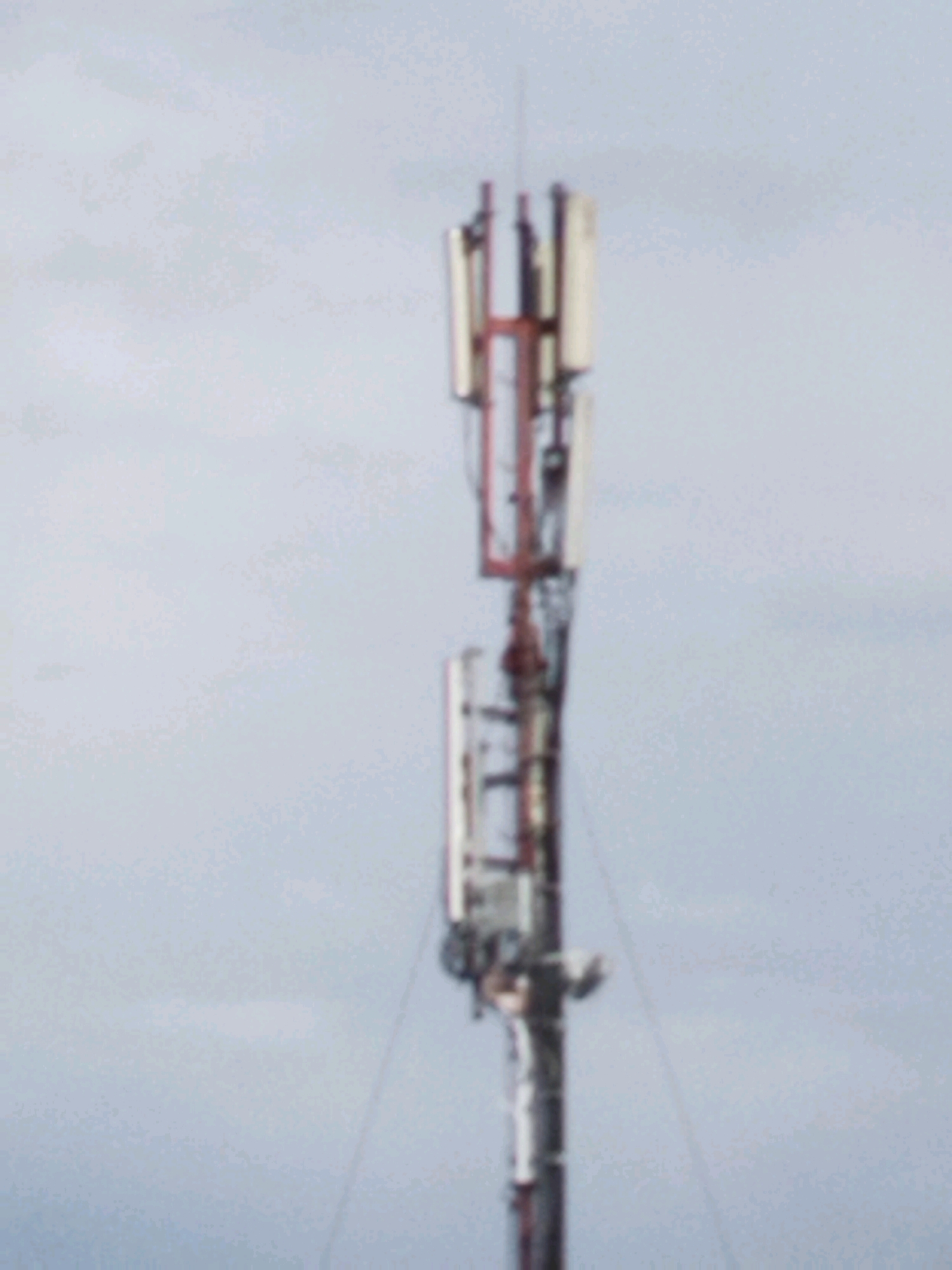 A cell tower stands against a hazy, overcast sky. The tower is a lattice structure with multiple antennas mounted at different heights. The antennas appear to be rectangular panels, some in white or silver, others with red trim. There are also various supporting structures and equipment visible on the tower. The image quality is poor, with significant blur and low resolution, making it difficult to discern fine details. The overall scene is devoid of any human presence or activity. The location is identified as Hrodna, Belarus.