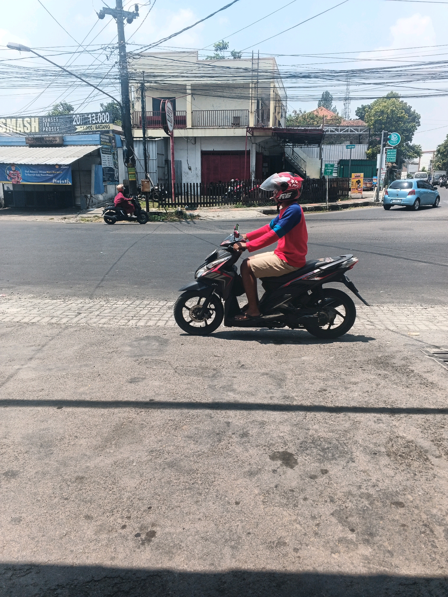 The image depicts a street scene in Ungaran, Indonesia, on a sunny day with a clear blue sky. In the foreground, a person is riding a black motorcycle. The rider is wearing a red and blue long-sleeved shirt, beige shorts, flip-flops, and a red motorcycle helmet with white and black accents. The motorcycle has red and black detailing.
In the background, there are several buildings and a significant amount of overhead electrical wiring. One building has a sign with Indonesian text, including "SMASH ITU PROSES" and prices like "200.000/BATANG". Another building displays a banner with an image of a person in a red and blue outfit, likely related to marketing or promotion. A blue car is visible on the road in the distance. The street is paved, with a sidewalk in the foreground that has some dark stains. The overall environment suggests an urban or semi-urban setting with active street life.