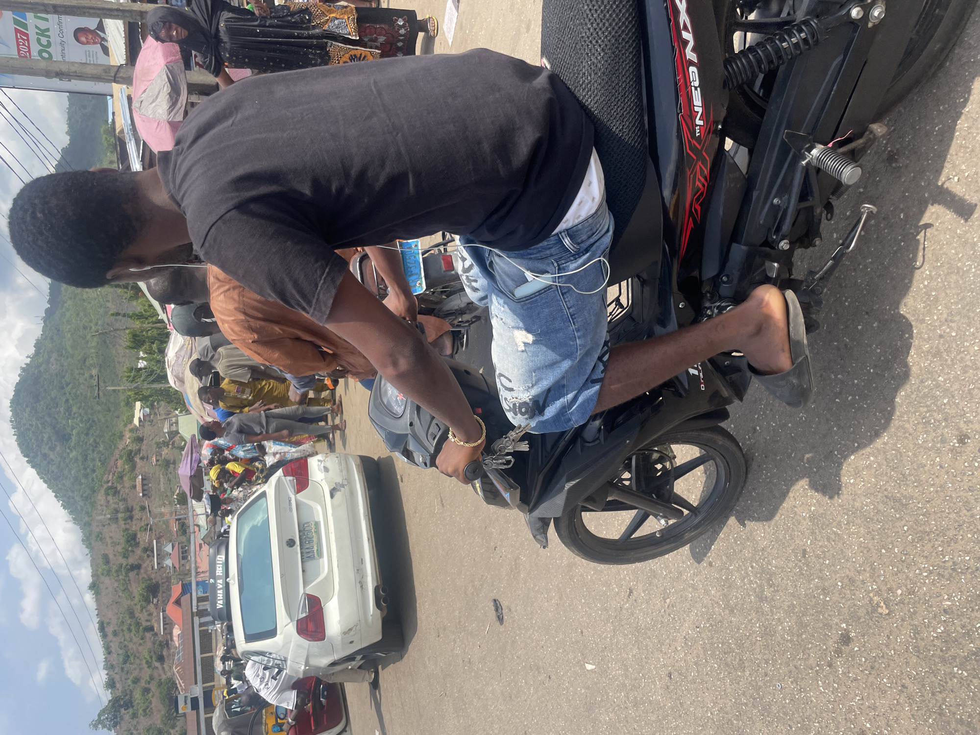 A medium-angle, outdoor daytime shot depicts a man seated on a black and red motorcycle on a street in Lokoja, Nigeria. He is wearing a black t-shirt, ripped blue jean shorts, and grey slip-on shoes. A gold-colored bracelet adorns his wrist. The motorcycle is parked near a white BMW car with the license plate "AAA 825 CS." Numerous people are on the street. In the background, there is a lush green mountain or hill. Buildings and houses are also visible in the background.