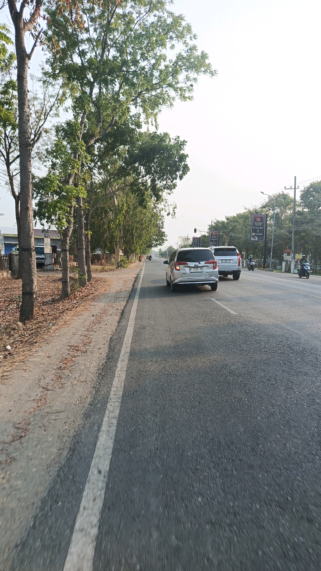 The image depicts a street scene in Babadan, Indonesia. A silver car with license plate N 3043 RS is traveling on the right side of the asphalt road, followed by another silver car. Two motorcycles are also visible on the road, one behind the silver cars and another further ahead on the right. The roadside features a row of trees with green foliage, and dry leaves are scattered on the dirt shoulder. A white line marks the edge of the asphalt road. In the background, there are buildings and utility poles. The sky is bright and clear, suggesting it is daytime.