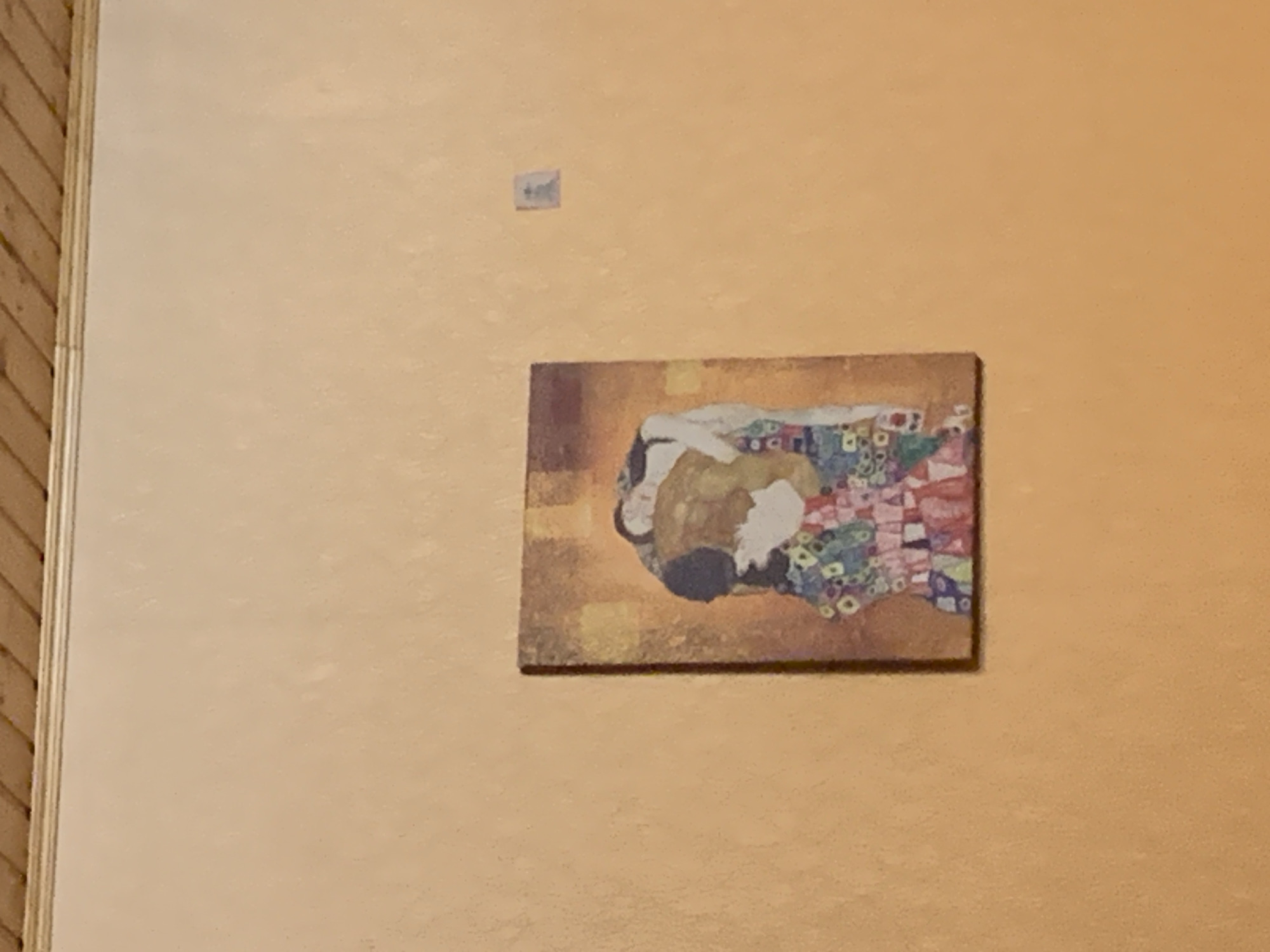 The image shows a framed painting of "The Kiss" by Gustav Klimt hanging on an orange-toned wall. The painting depicts a couple embracing in a colorful, patterned robe. To the right of the painting, a small, square blue and white sticker is affixed to the wall. Above the wall, a wooden slatted ceiling is visible, with a decorative trim at the intersection of the wall and ceiling. The painting is the primary subject, with the sticker and ceiling serving as contextual elements within an indoor environment.