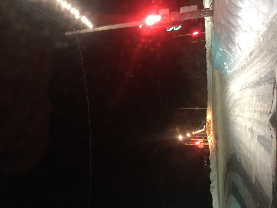 The image displays a nighttime winter street scene in the city of Kremyonki, Russia, viewed from a low perspective, likely from inside a vehicle. The foreground reveals a snow-covered asphalt road with distinct tire tracks, flanked by elevated snowbanks. To the right, a utility pole stands prominently, supporting a traffic light emitting a bright red signal. A fainter green signal is visible beneath the red light. A string of luminous white lights is wrapped around the upper section of this utility pole, extending vertically. Another red traffic light is observable further down the road on the left. The road continues into the distance, lined by a series of streetlights on utility poles that cast an amber glow on the snowy environment. The background is dominated by a dark, featureless night sky, with subtle, distant outlines of terrain or structures visible on the horizon. A thin wire traverses horizontally across the upper-mid portion of the frame. No people or active interactions are depicted.