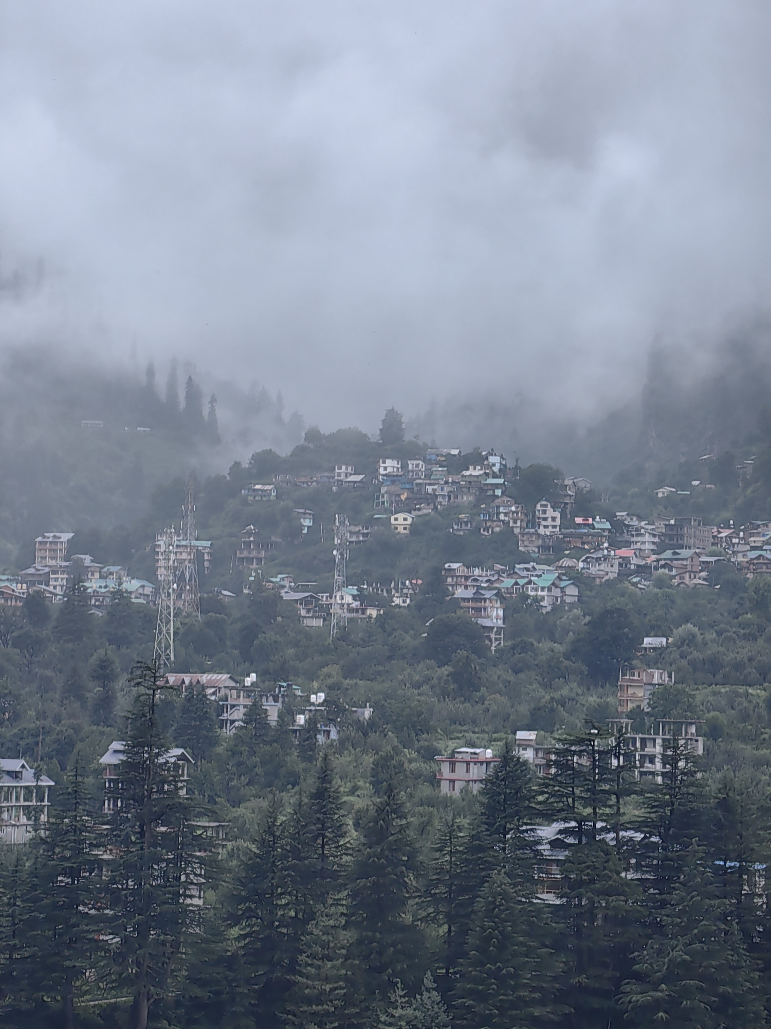 The image depicts a dense, overcast mountain village in Manali, India. Numerous buildings, ranging from multi-story structures to smaller houses, are scattered across the steep, tree-covered slopes. Many of the buildings have distinctively sloped, often green or red, roofs. Tall, slender communication towers are prominent in the mid-ground, interspersed among the buildings. Dense evergreen trees, likely pine or deodar, dominate the landscape, particularly in the foreground and between structures. The overall atmosphere is misty and subdued, with clouds and fog obscuring the upper reaches of the mountains and lending a somewhat hazy quality to the scene. No people are clearly visible in the image. The environment suggests a high-altitude, densely populated area within a mountainous region.