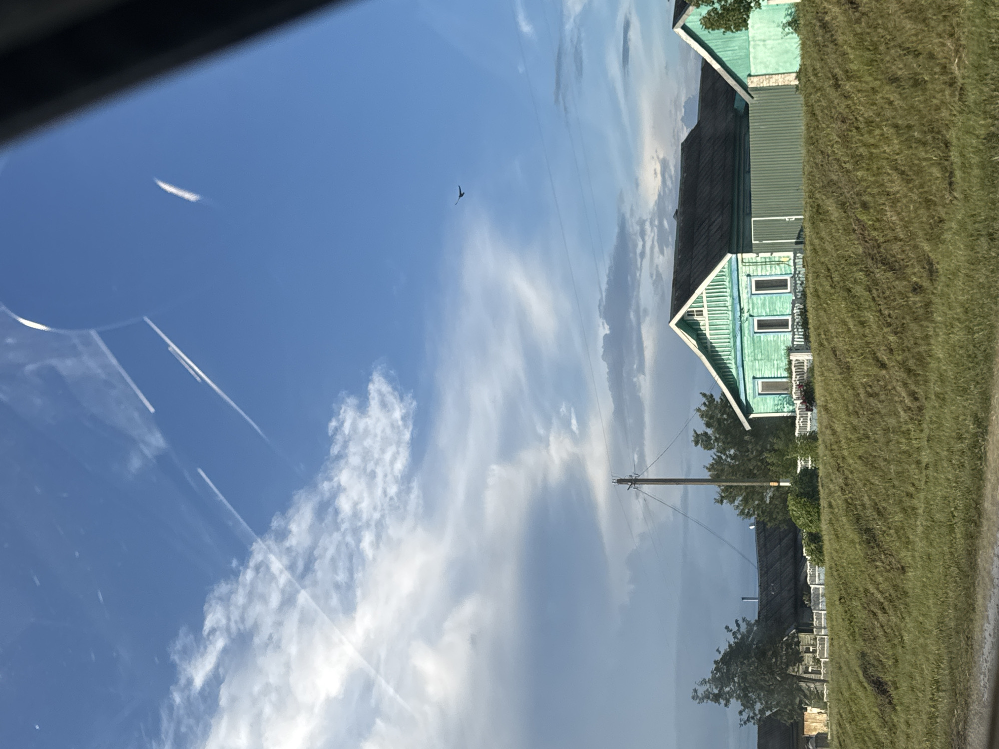 The image captures a pastoral scene under a bright, blue sky with wispy white clouds. A thin, contrail from an aircraft streaks across the sky on the left side of the frame. In the distance, a bird is seen in flight. The scene is dominated by a row of traditional green wooden houses with dark roofs, typical of rural Belarus, within the Акцябрскі сельскі Савет region. One house in the foreground displays characteristic white trim around its windows and roofline, with multiple windows visible on its facade. The houses are bordered by a lush, green grassy embankment on the right, suggesting a roadside setting. A utility pole with wires is also visible, indicating the presence of infrastructure in this rural area. The bright sunlight and clear sky suggest it is daytime, likely late morning or early afternoon. The overall impression is one of a serene and tranquil rural landscape. There is no visible text in the image.