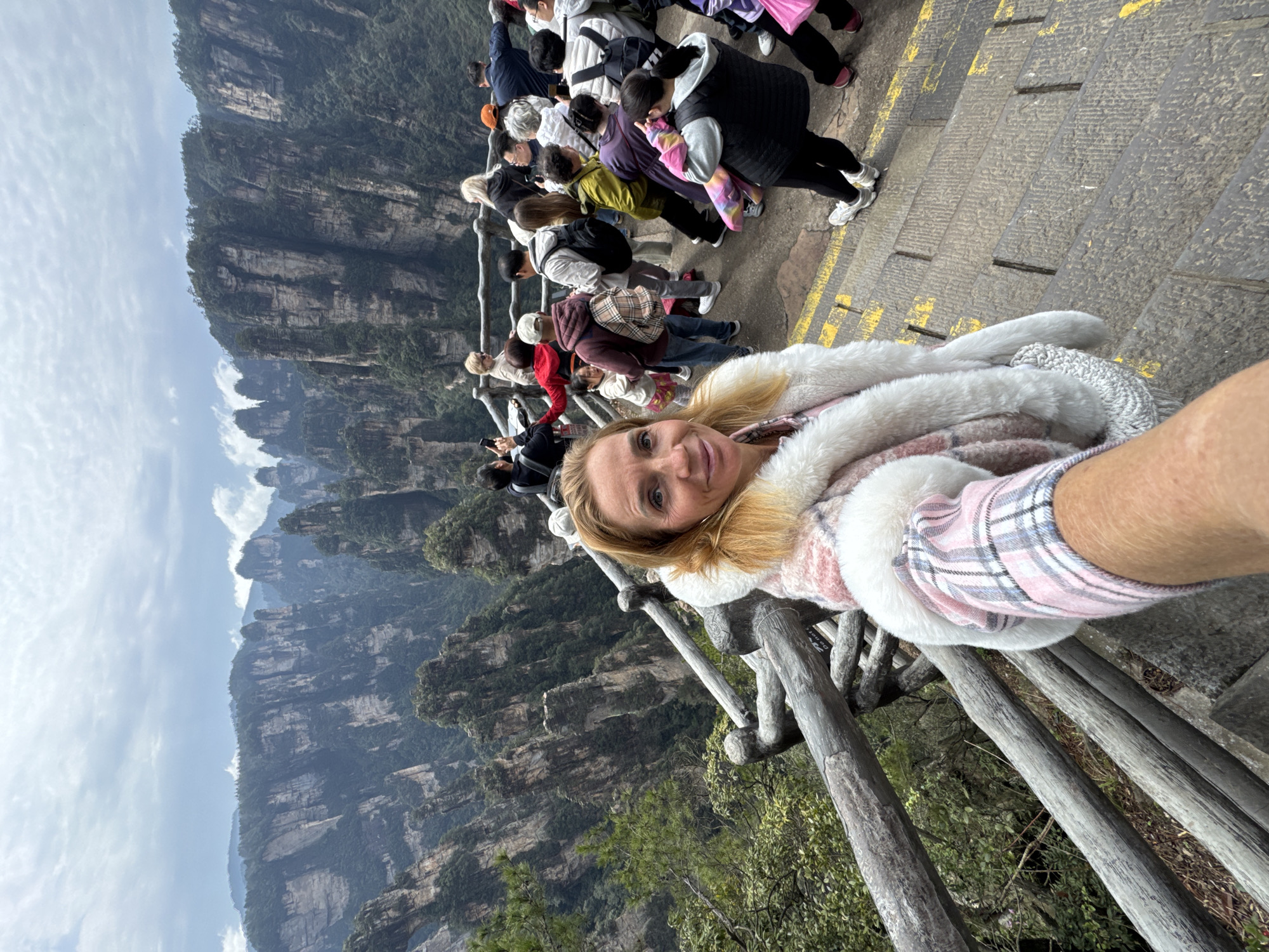 The image depicts a woman taking a selfie on an outdoor viewing platform within Zhangjiajie National Forest Park, Wulingyuan Scenic Area, which is associated with the broader region of Luoguta, China.
In the immediate foreground, a fair-skinned woman with shoulder-length blonde hair is visible from the chest up, smiling directly into the camera. She wears a pink and white plaid long-sleeved shirt under a thick white faux fur vest. Her right arm, tanned, is extended, holding the camera for the selfie.
To the left of the woman, a weathered wooden railing with horizontal logs and vertical posts serves as a safety barrier. Beyond the railing, dense green foliage and the steep drop-off of the mountain are visible.
Behind the woman, a crowded, paved stone staircase descends. Numerous individuals of various ages and ethnicities are present on the stairs and platform. Most are dressed in casual outdoor attire, including jackets, hoodies, and backpacks, consistent with hiking or touring. Many are looking out towards the panoramic view, some are holding smartphones, seemingly taking photos or videos. A few individuals are leaning on a similar wooden railing that extends along the viewing area.
The background reveals the distinctive and dramatic landscape of Zhangjiajie, characterized by towering, pillar-like sandstone peaks covered in dense green vegetation. The sky above is overcast with a mix of light and dark grey clouds, indicating a cloudy day. The overall impression is that of a popular tourist destination with breathtaking natural scenery.